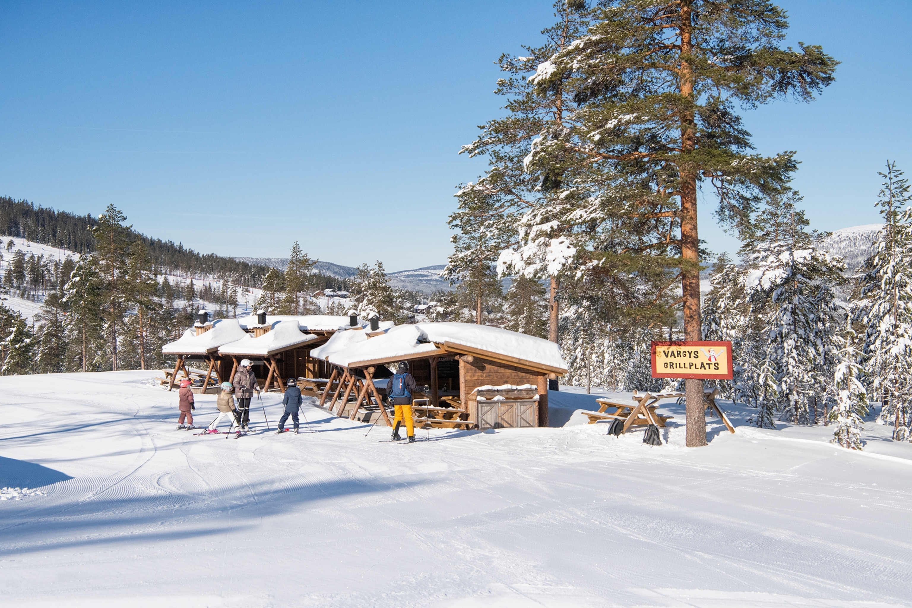 A cluster of snowed-in, simple wooden huts on the side of a piste with a group of skiers looking for a pit stop.