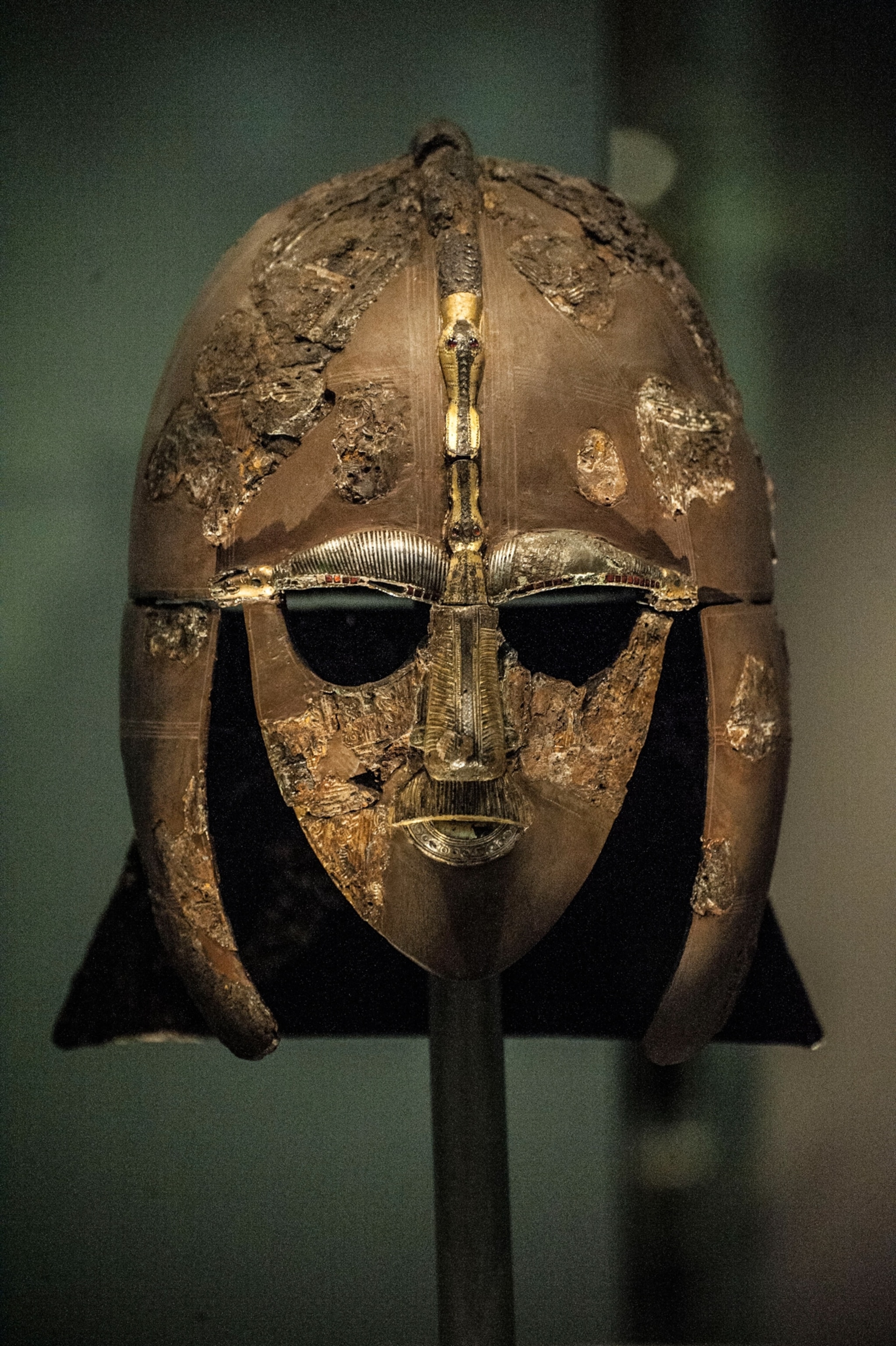 An ancient, weathered metal helmet featuring a decorative gold and silver face mask with intricate engravings, mounted on a stand against a dark background.