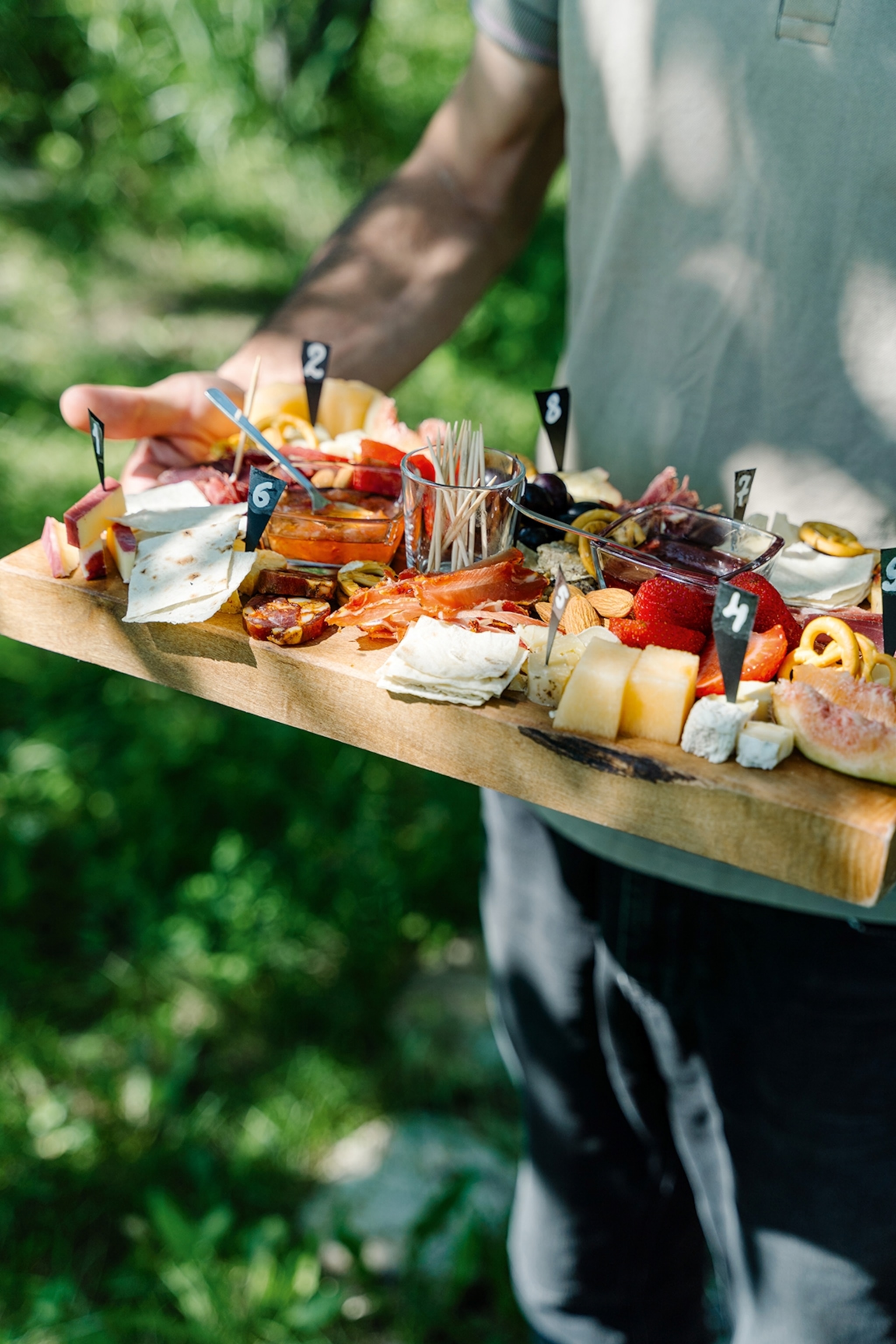 A person holding up a wooden charcuterie board with feathered out cheese slices, cured meats crisps and fruit.