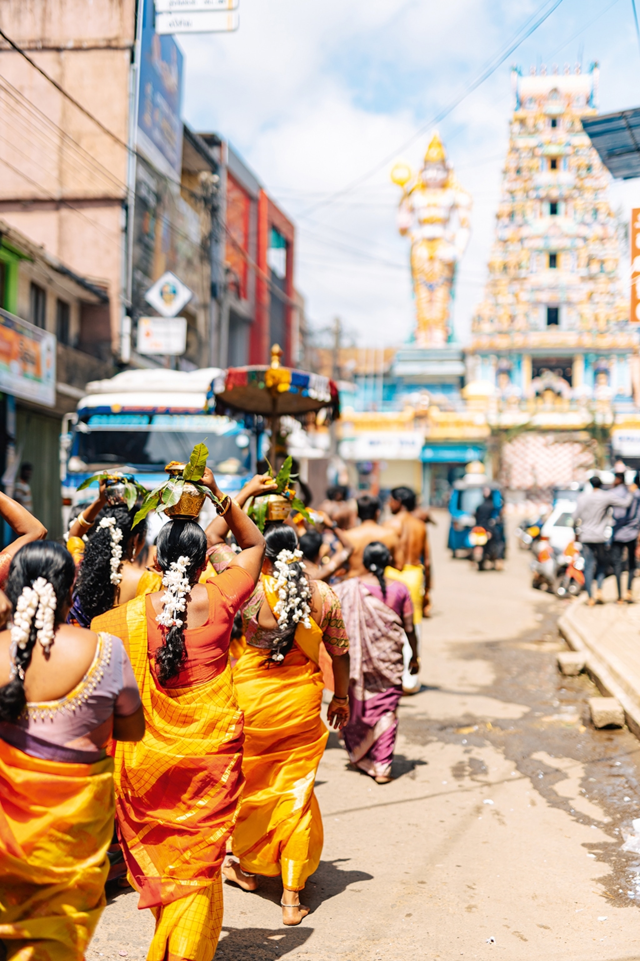 A busy city scene with a group of local woman shot from the back, carrying urns on their heads and walking towards a towering and playful-looking temple.