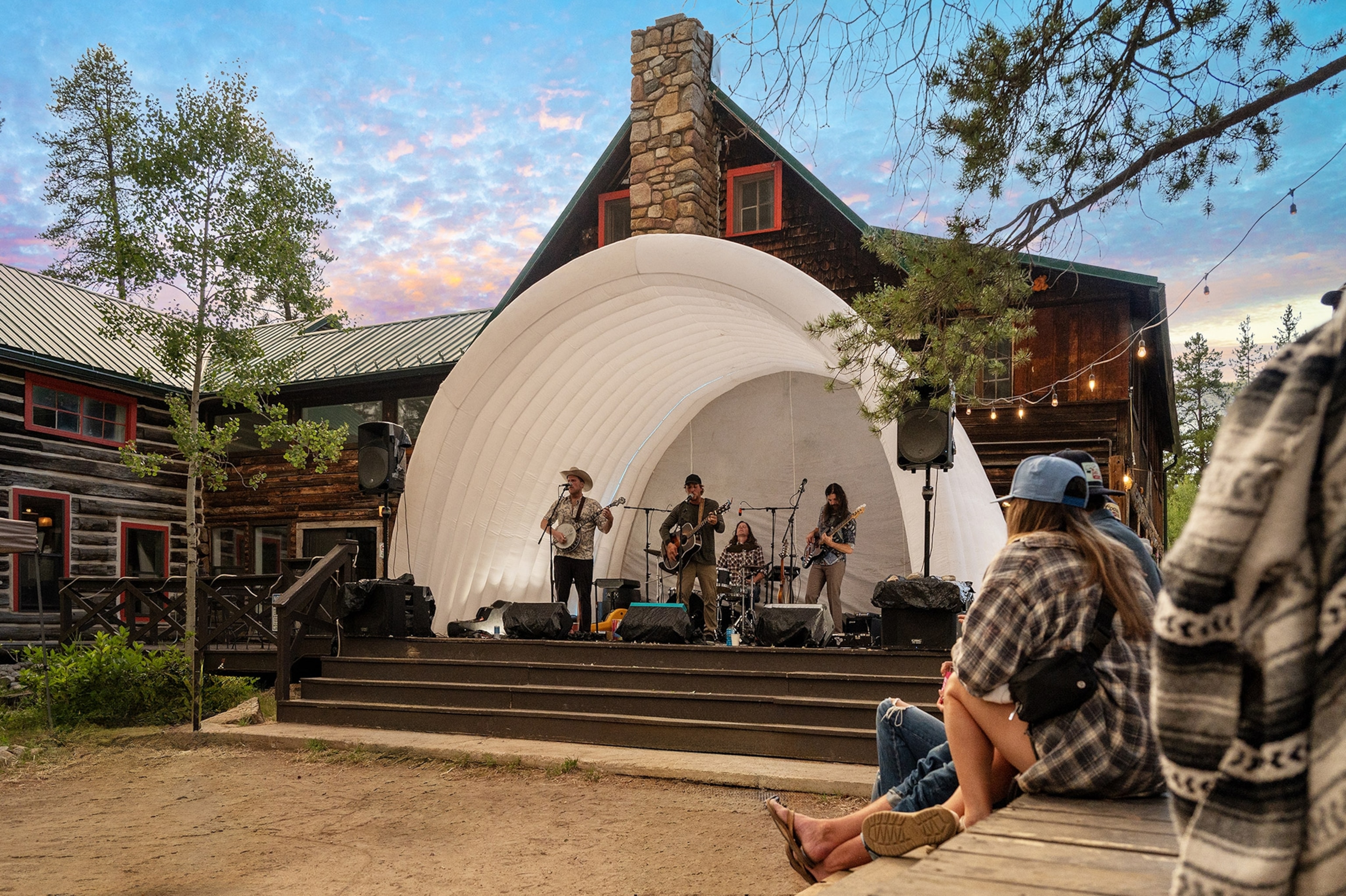 A small crowd gathers around an outdoor stage in front of a lodge, while a band performs