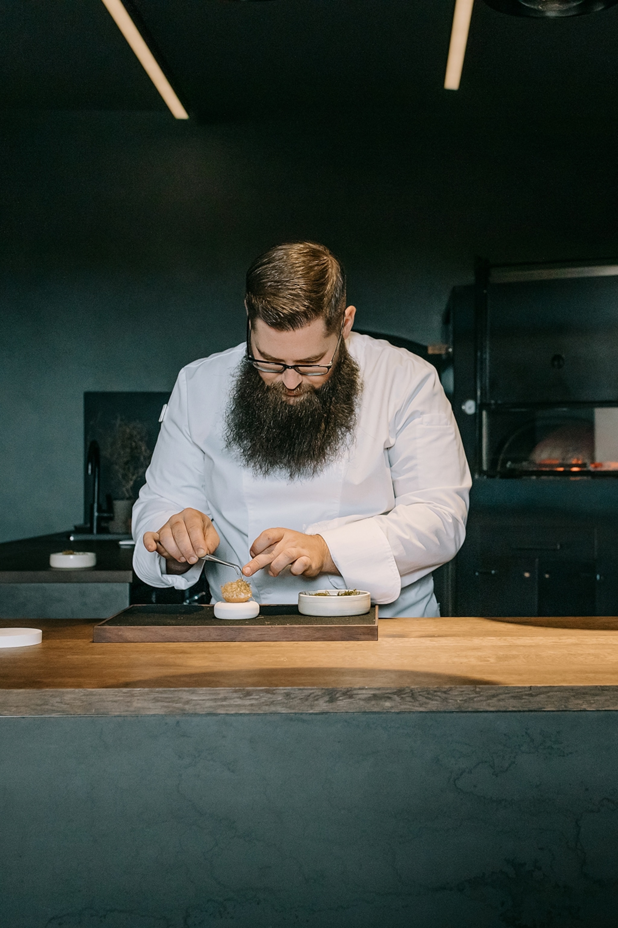 A young chef on a lit pass, adding final touches to a delicate dish.