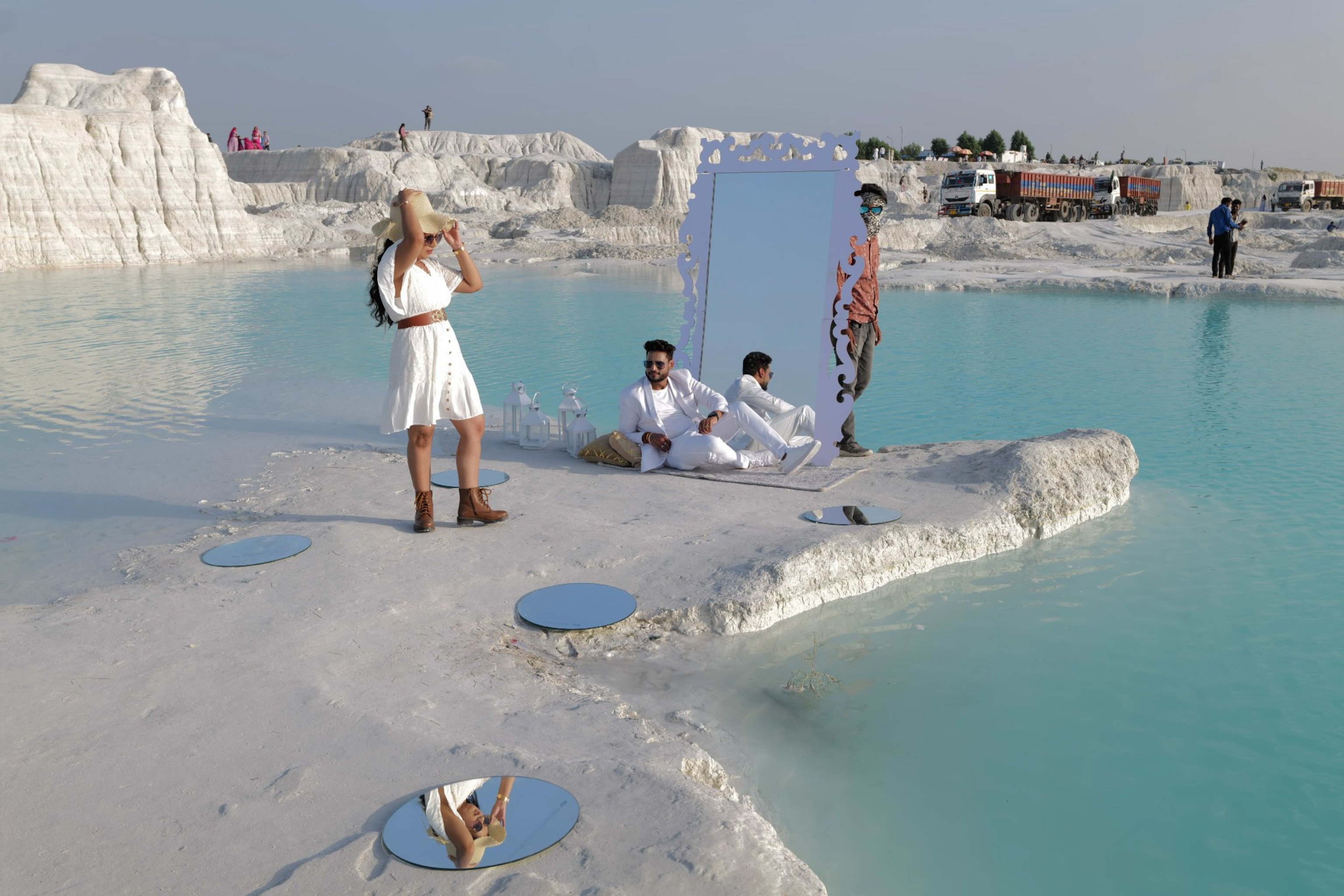 A woman holds the brim of her hat her reflection can be seen on the mirror on the floor, a man stares lovingly at her from the ground with a larger mirror behind him. Another with a face covering and shades peaks out behind the mirror cheekily. The white tones of the landscape are contrasted by bright blue water large brown trucks can be seen nearby.