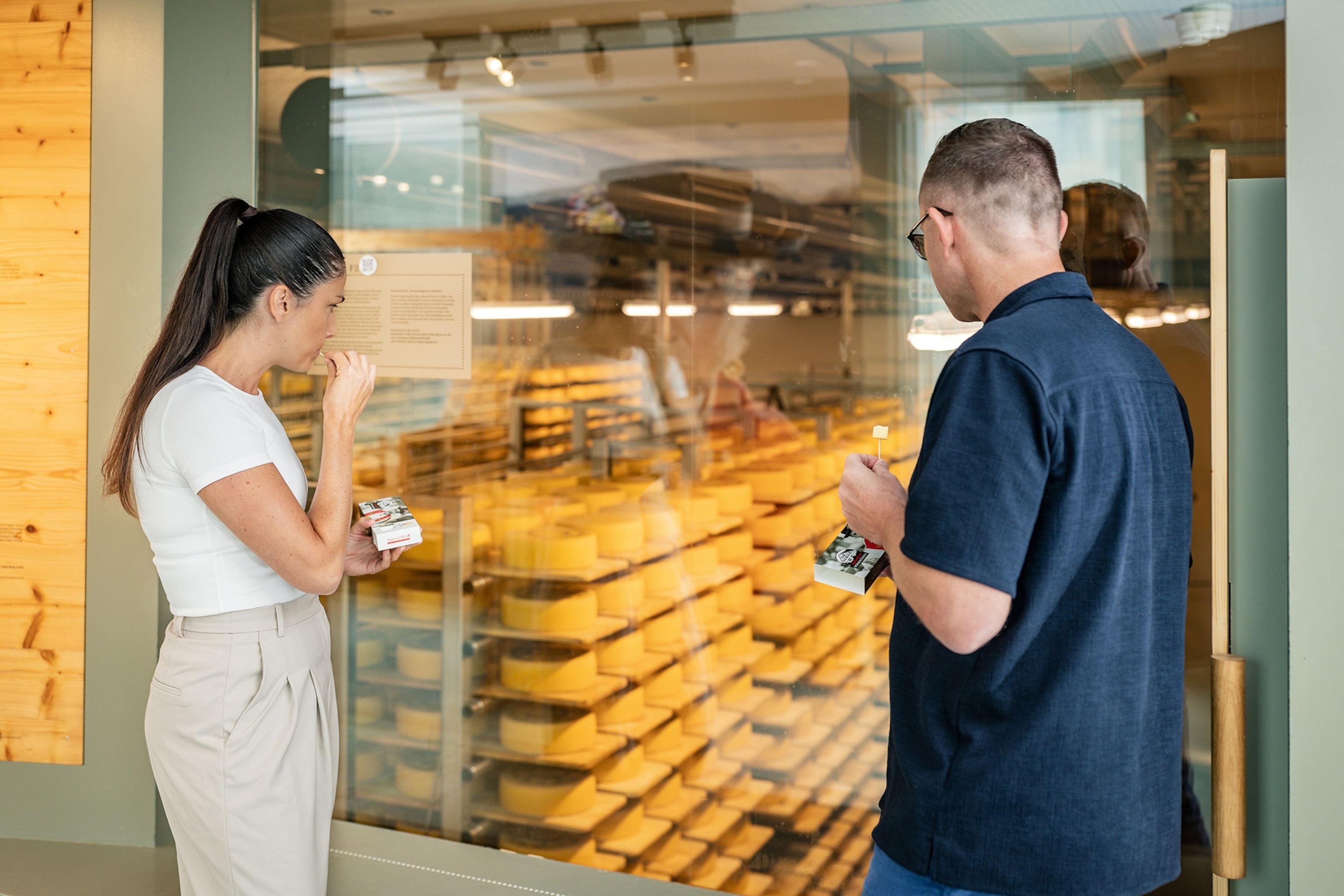 Two visitors standing in front of a glass window into a storage room with cheese.