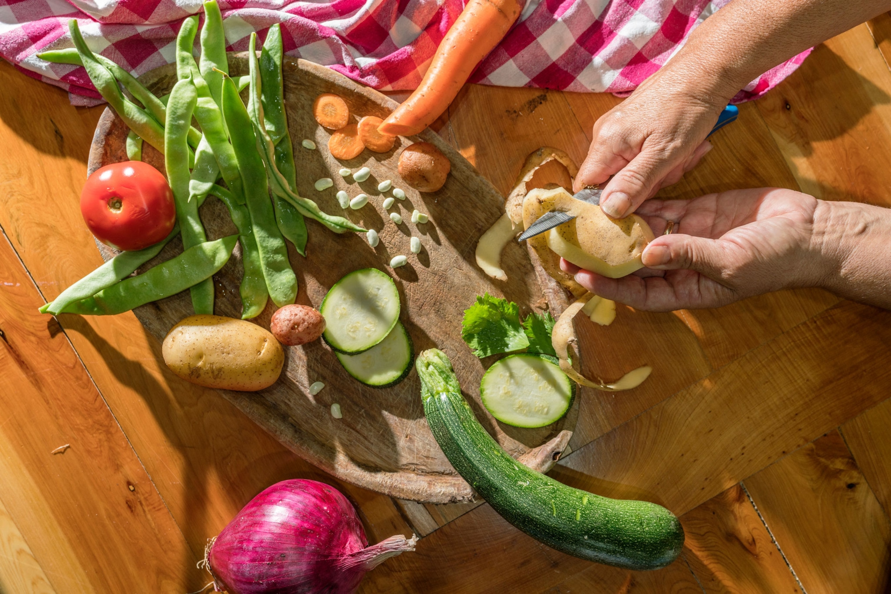 Hands peel a potato over a plate of vegetables.
