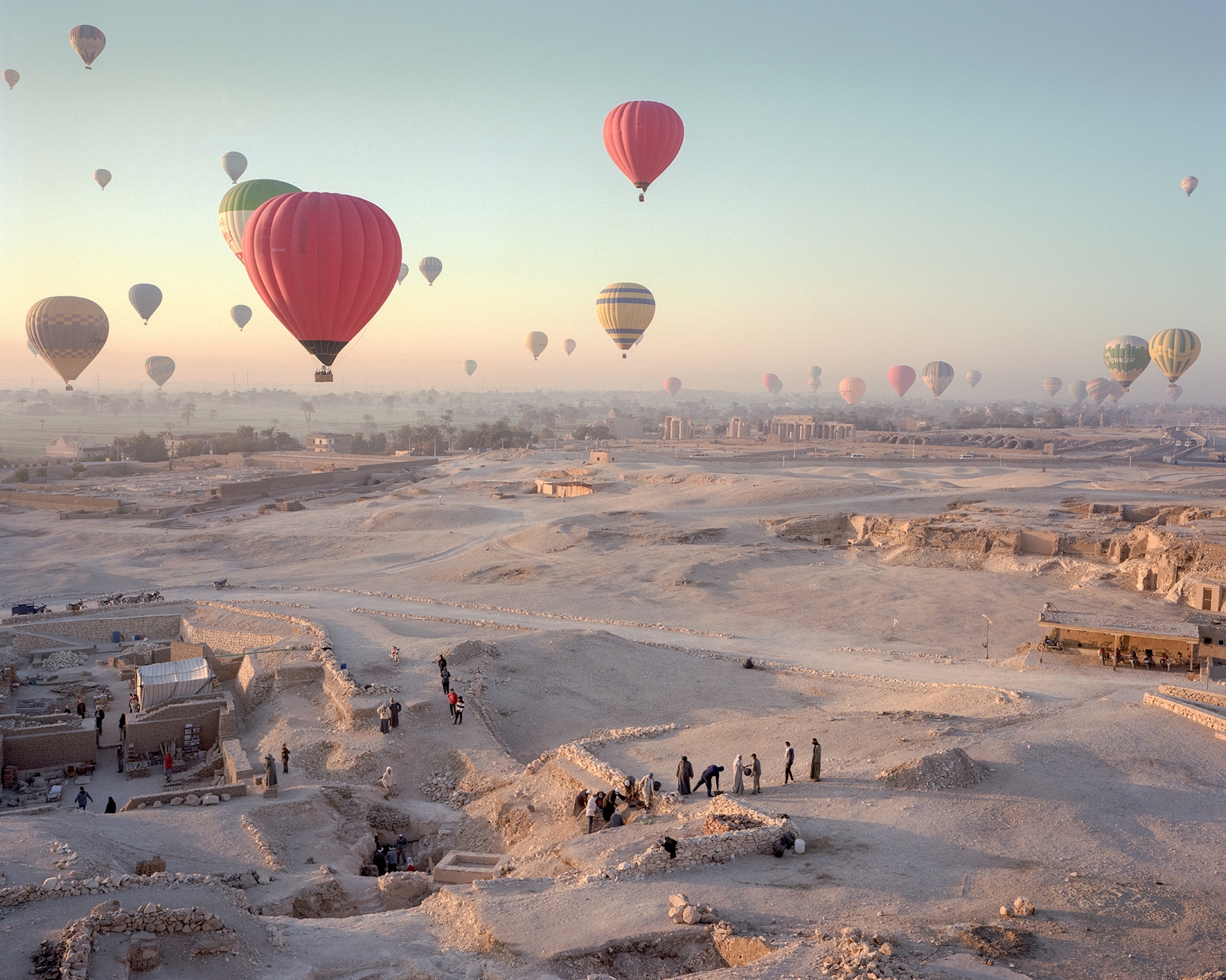 Hot air balloons float above an archaeological site in a desert landscape at sunrise, with excavation areas, ancient ruins, and small groups of people visible across the sandy terrain below.