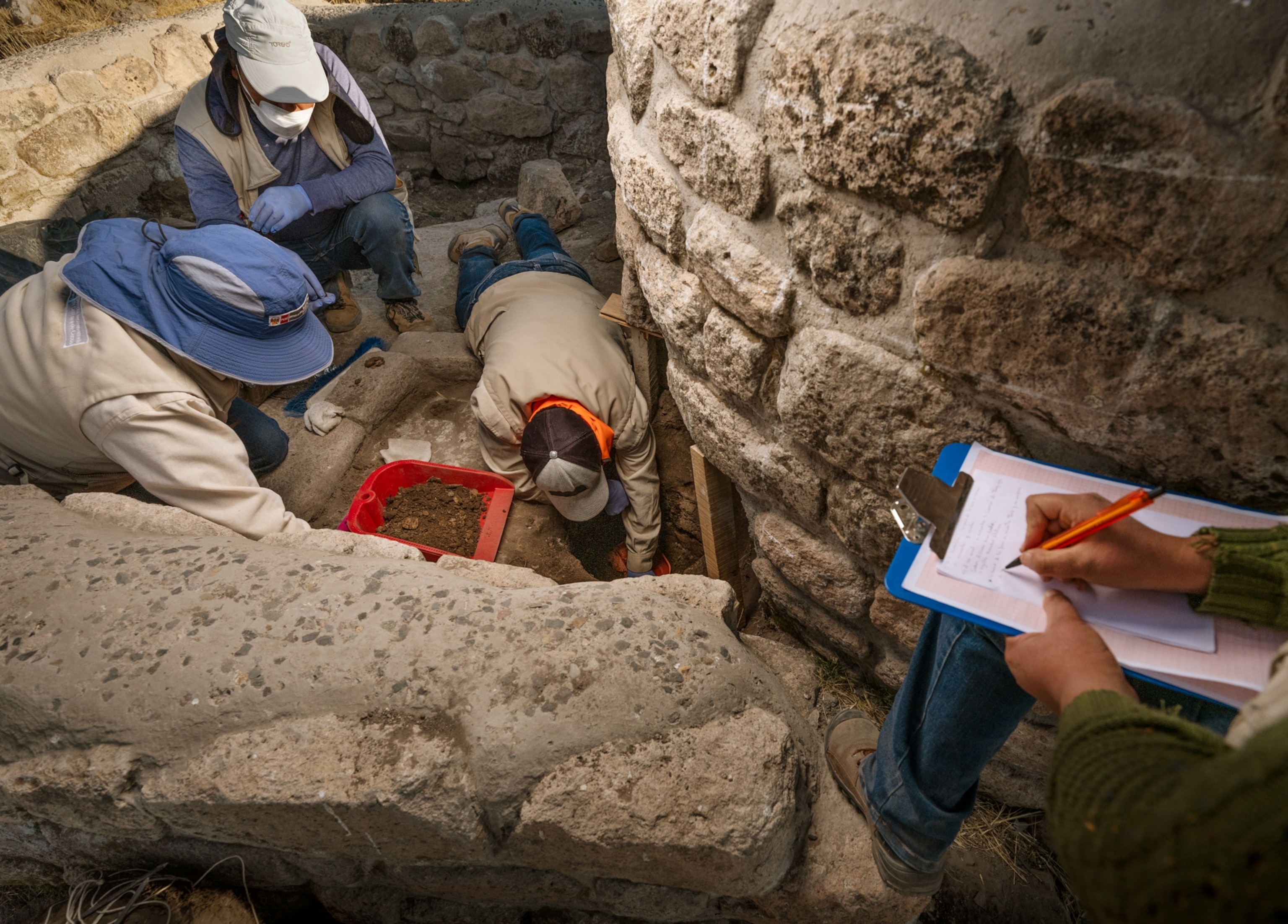 One person digging into soil by the stone wall, two people watching him, and another one is on the wall above them taking notes on the notepad.