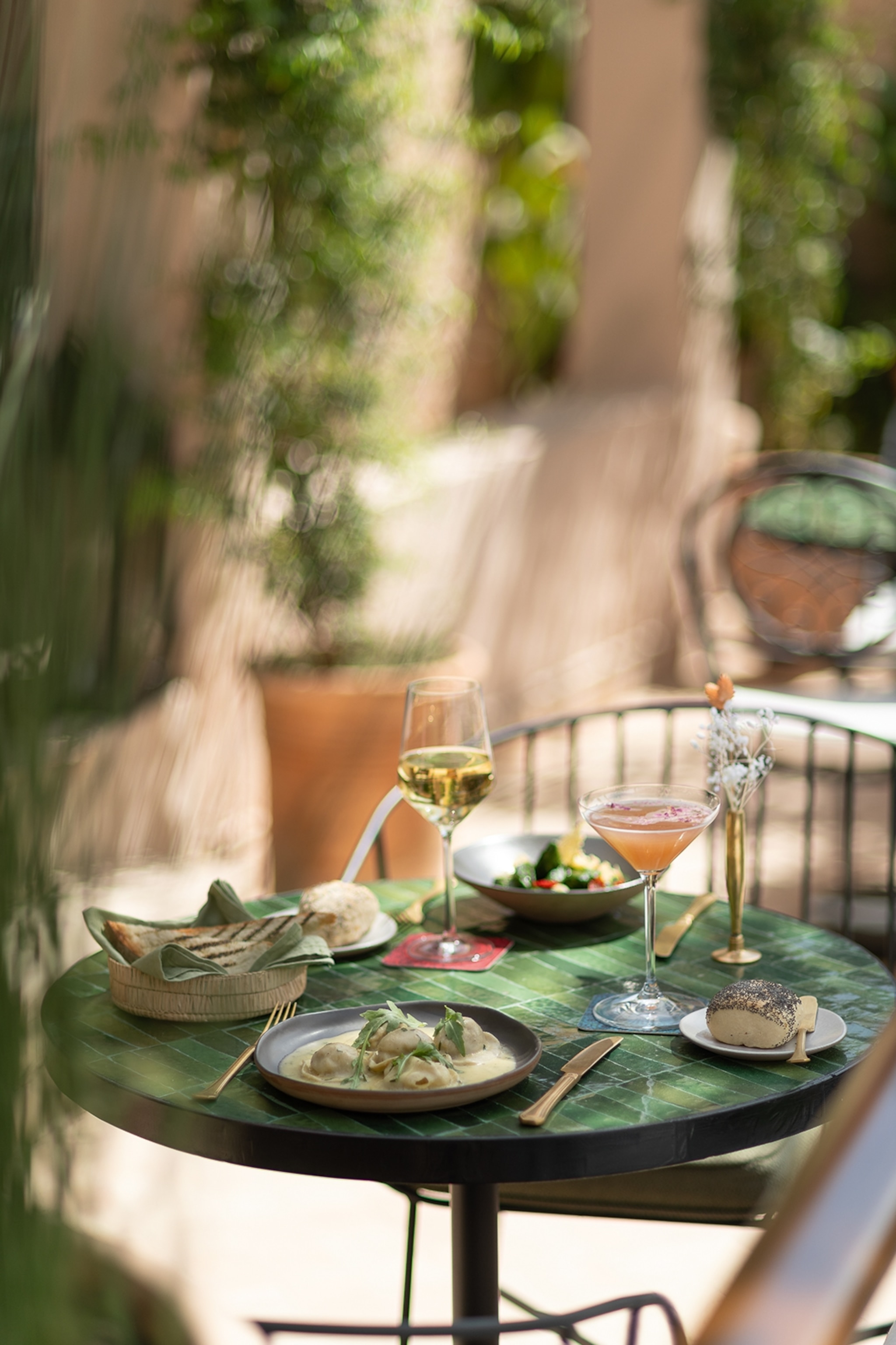 A close-up shot of a tiled bistro table featuring plates of food and cocktails in an out-of-focus garden.