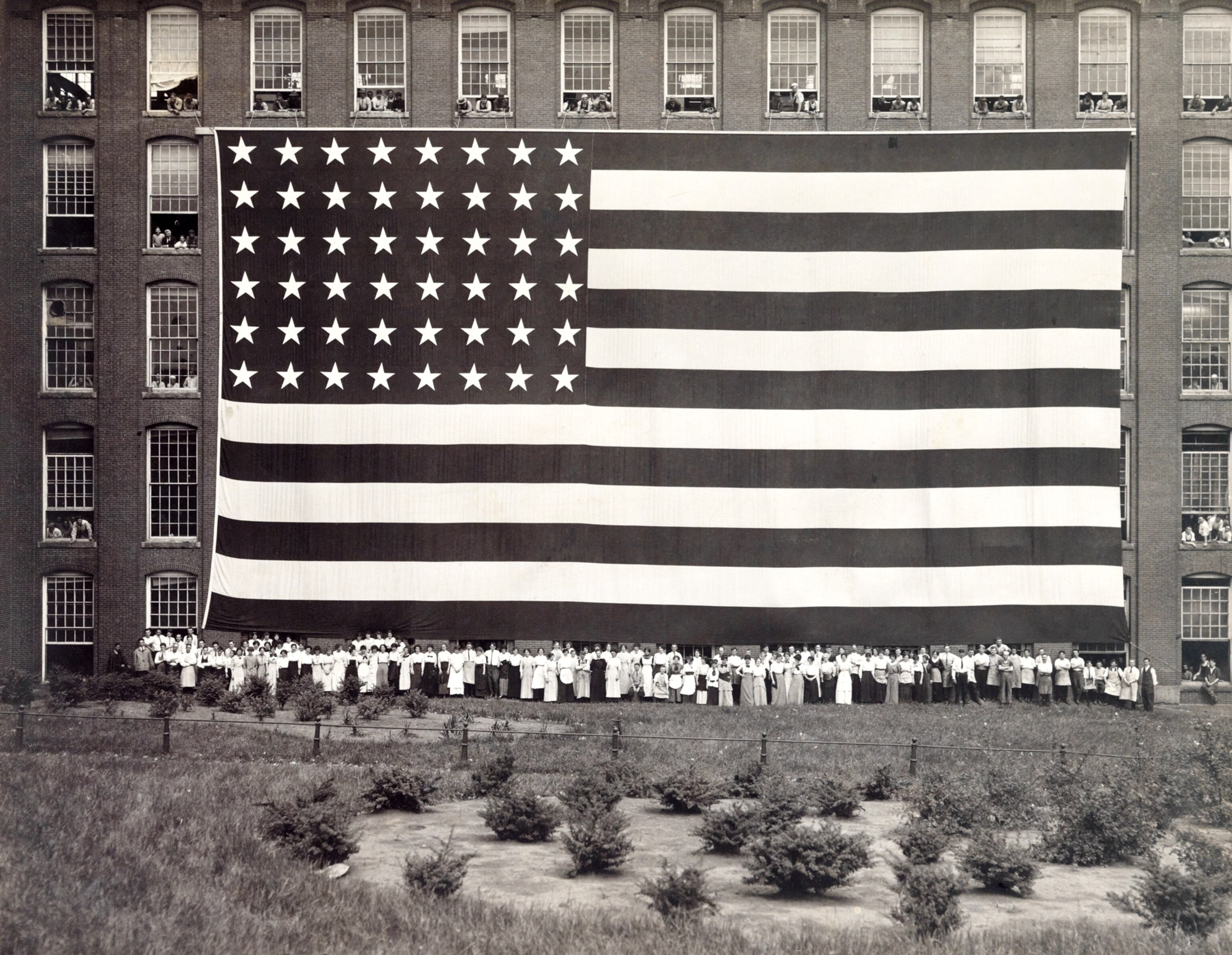 Dozens of people stand in front of a four-story American flag, 1910s.