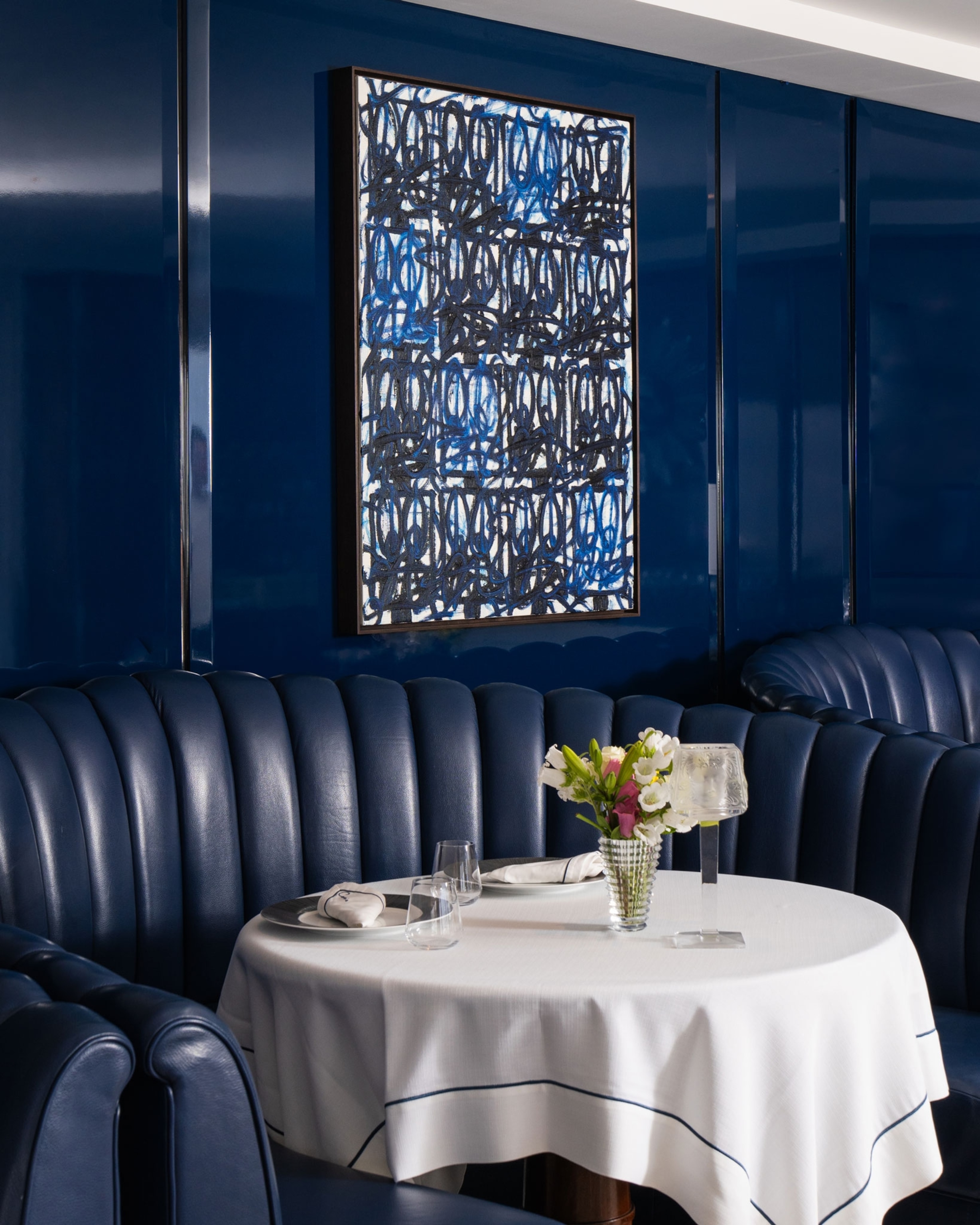 A blue, abstract painting hanging on a dark blue wall in a restaurant with dark blue upholstered booths, round tables and a white table cloth.