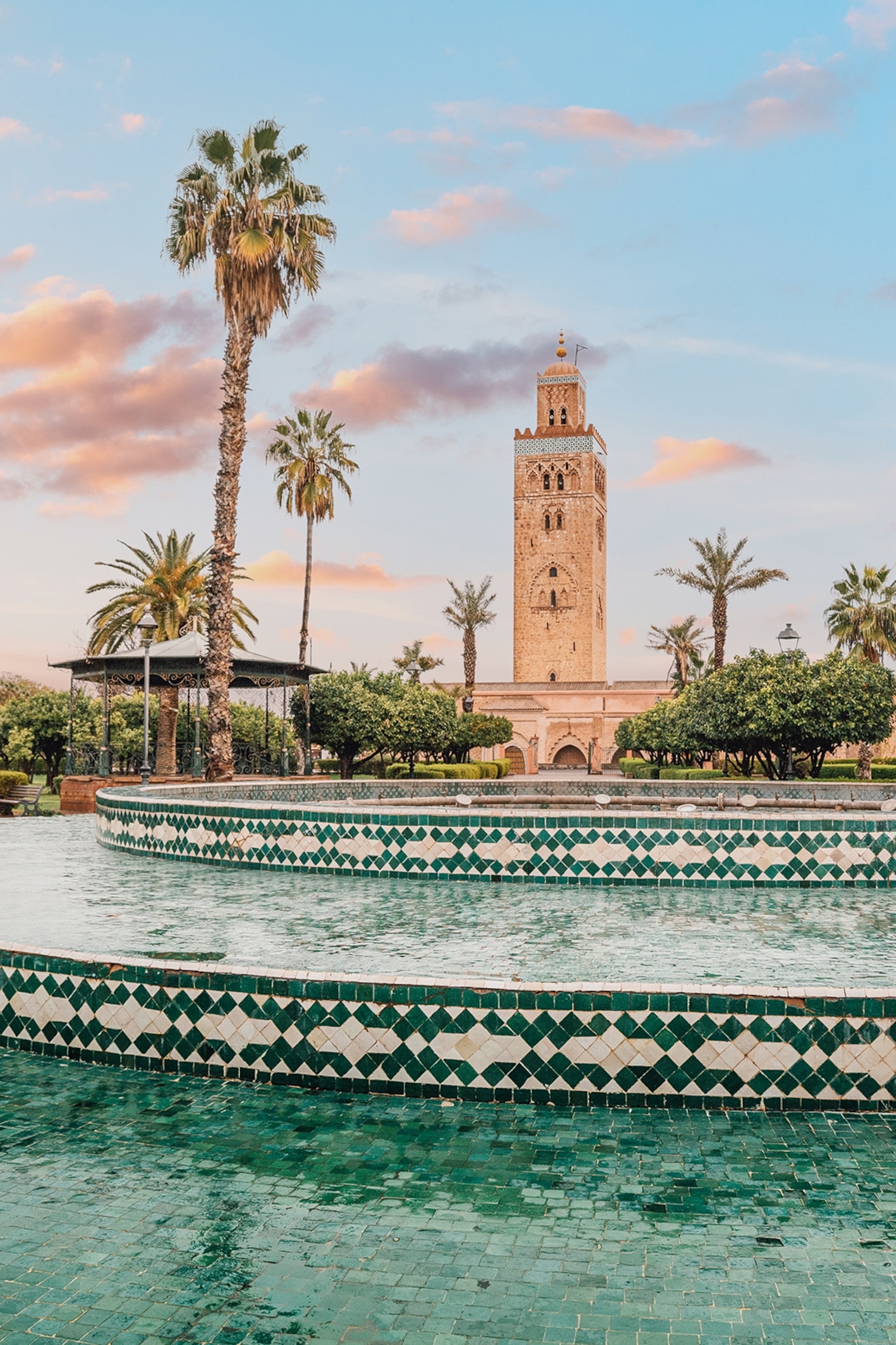 A wide and tiled fountain with palm trees and a stone mosque in the distance.