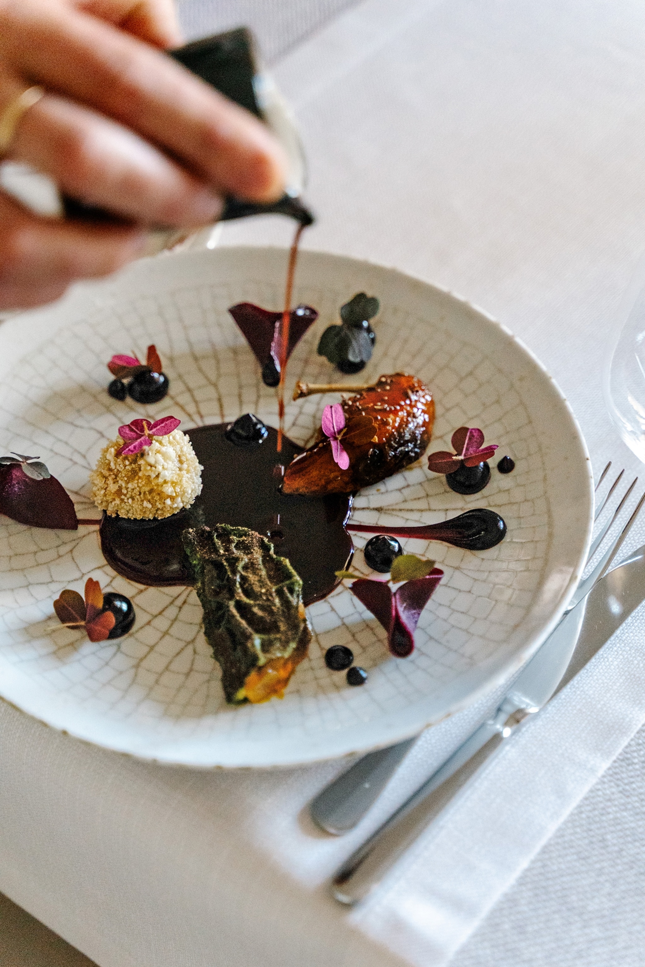 A hand pours a dark sauce over an artfully plated dish featuring vegetables, garnishes, edible flowers, and carefully arranged ingredients on a white ceramic plate.