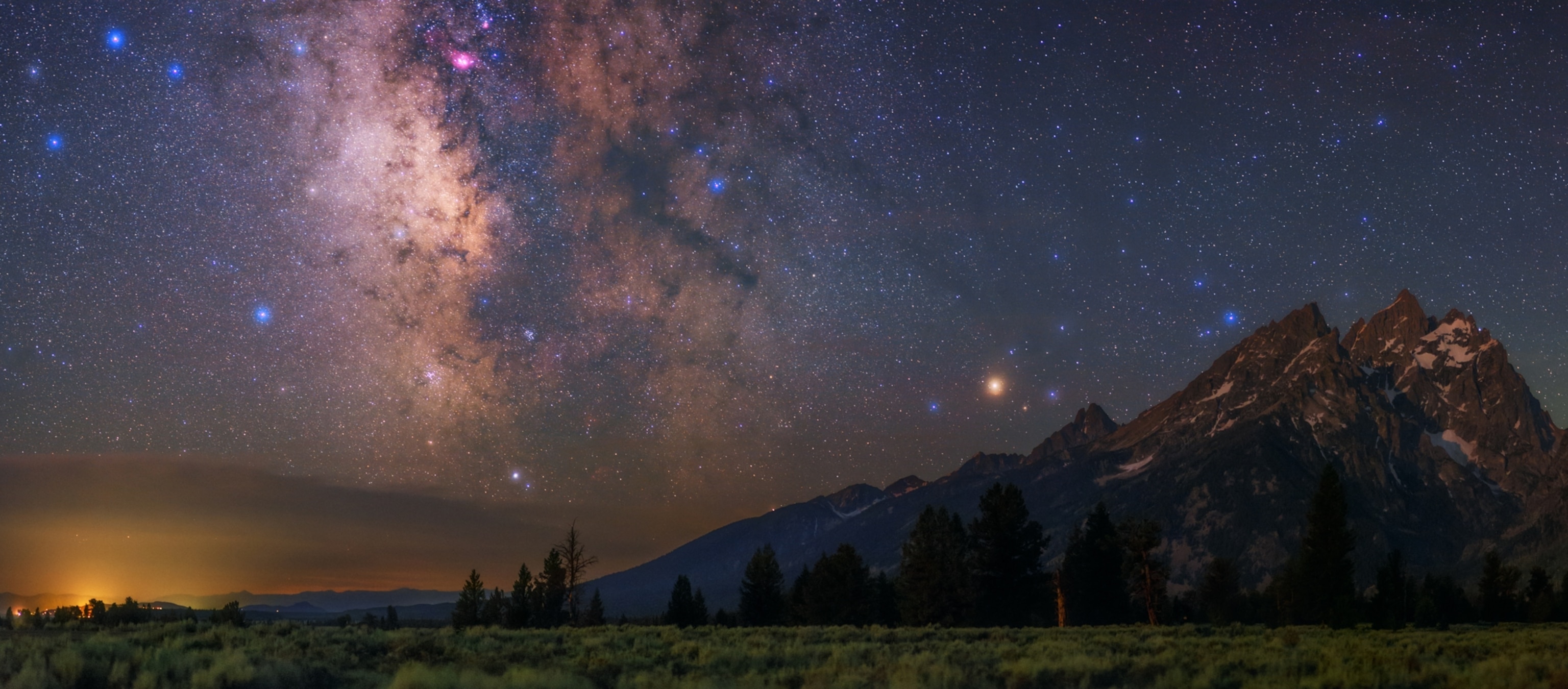 A starry sky with bright Milky Way over silhouetted mountains and forest