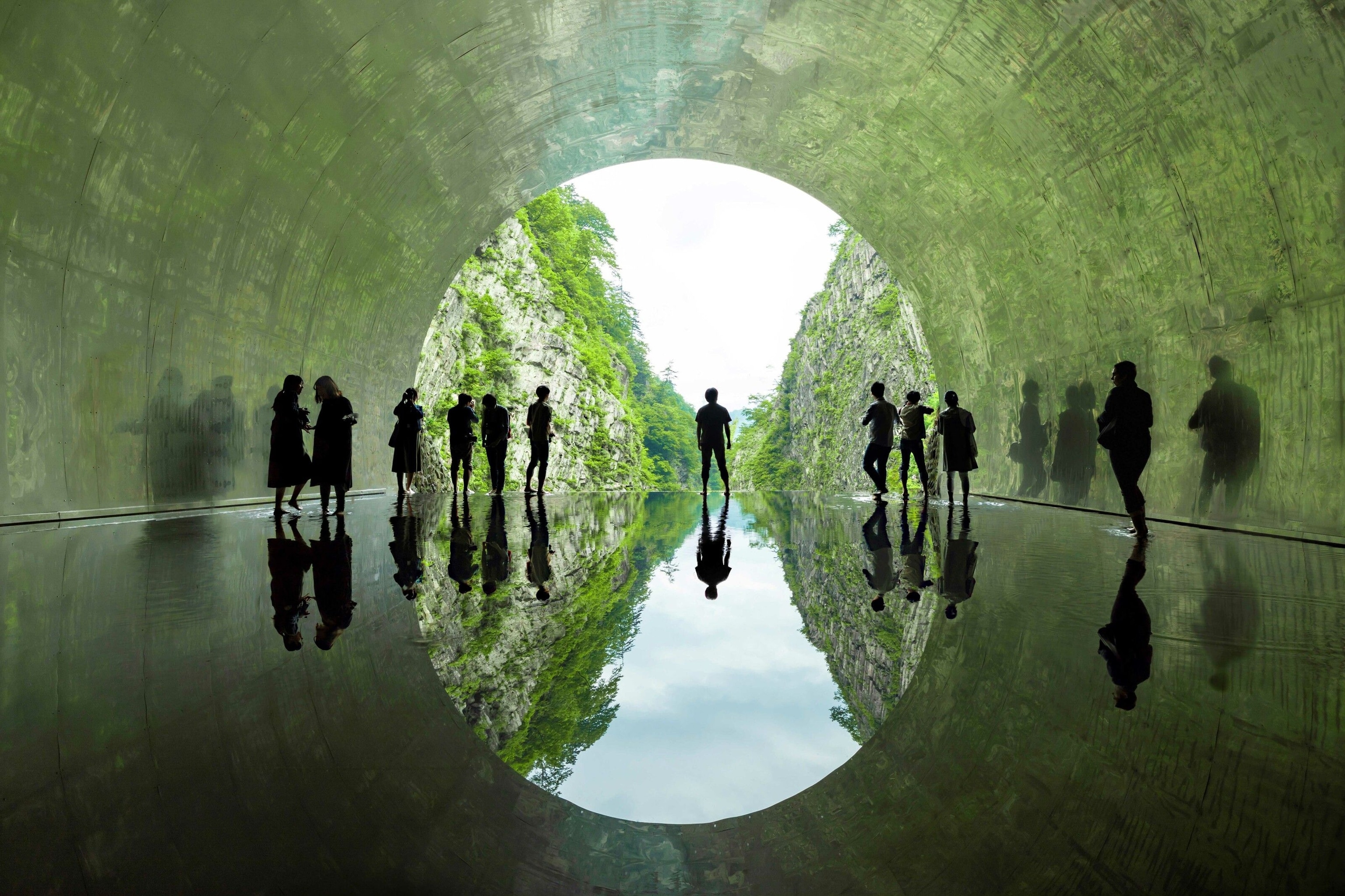 An artistic space. POV it looks like we are looking through the underside of a large concrete bridge, but the floor is a thin glassy layer of water, reflecting the outside and the people stood on it.