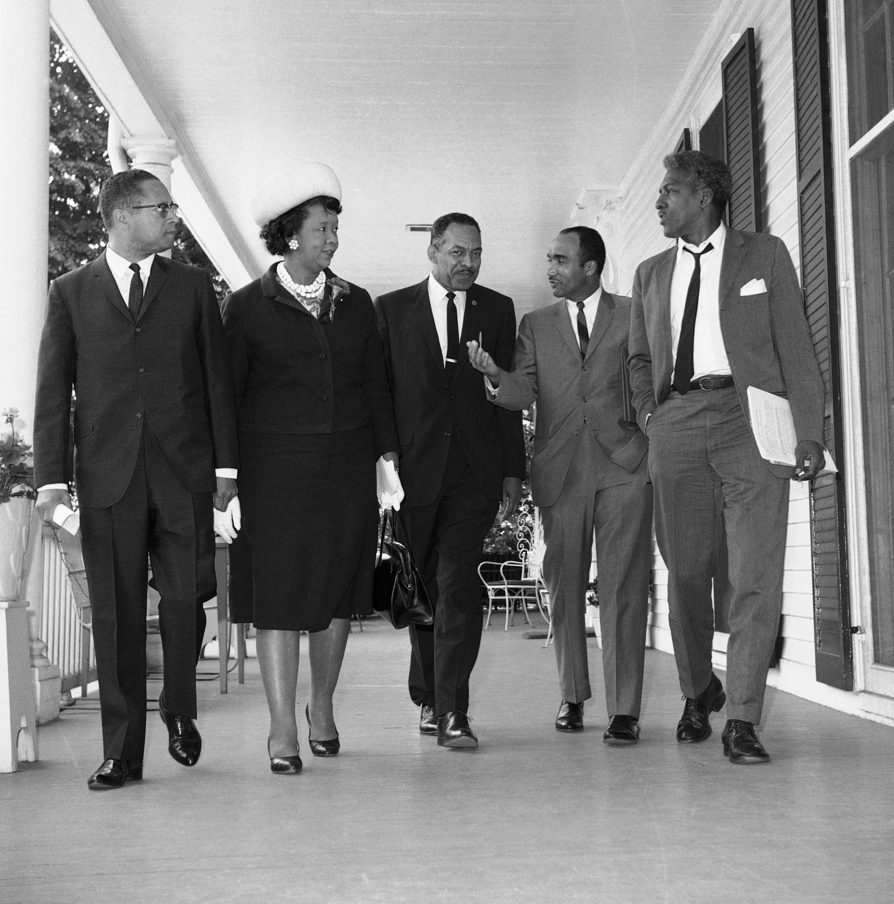 A group of five people, four men and one woman in formal attire, walk together on a porch.