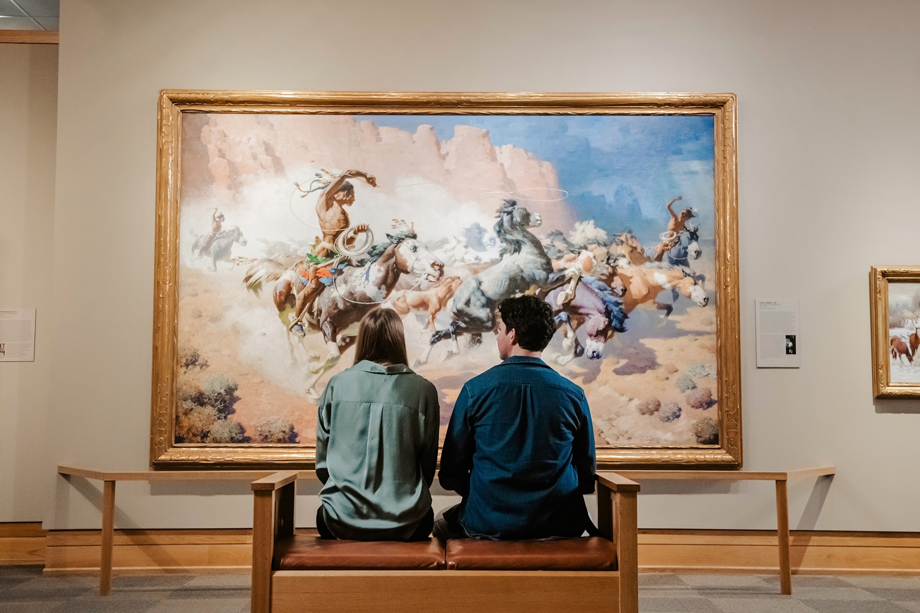 A man and woman sit on a bench admiring a painting of men on horseback.