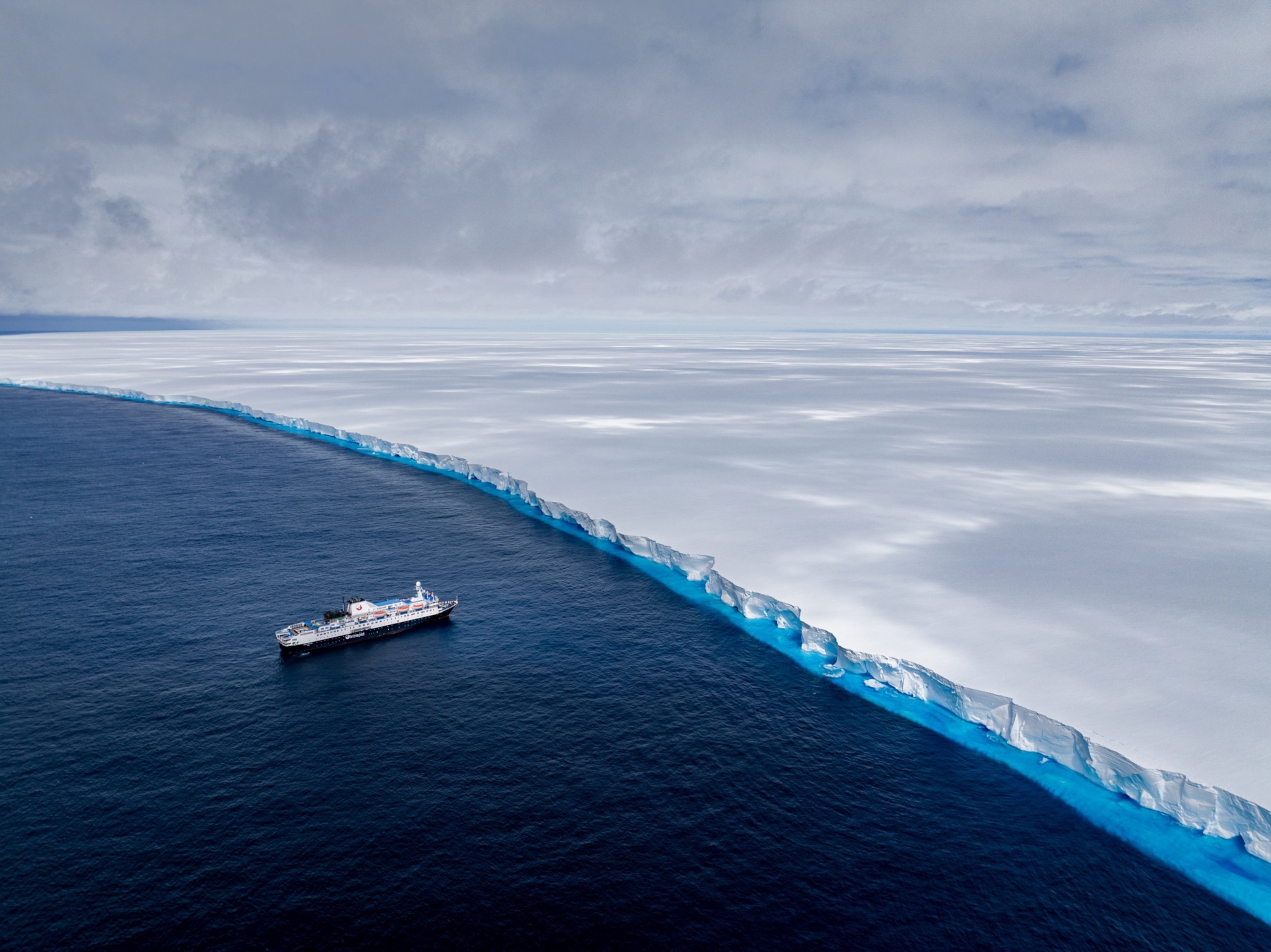 A ship sails along a vast, jagged edge of an iceberg under a cloudy sky