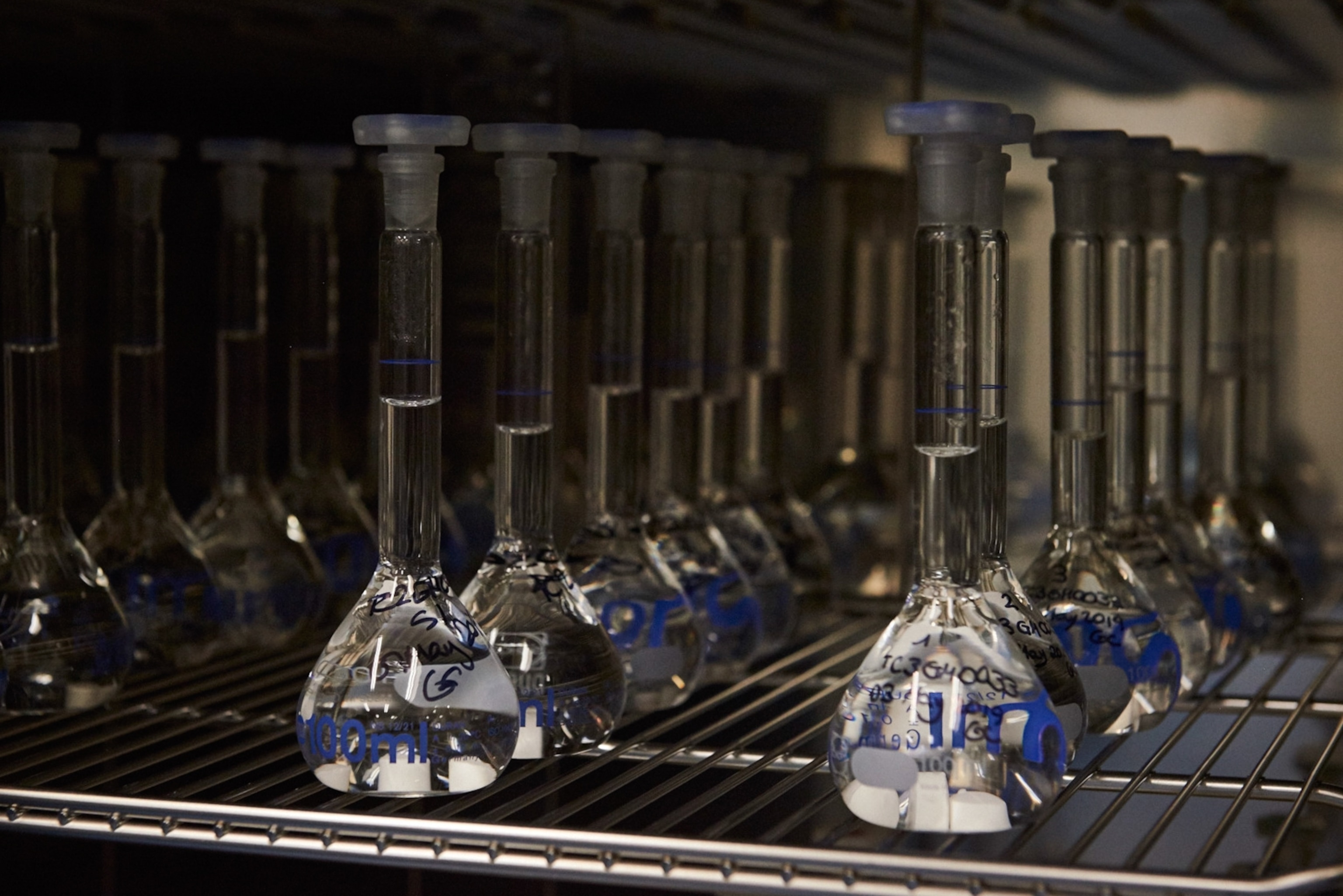 Rows of clear glass volumetric flasks with liquid and stoppers sit on a metal rack.