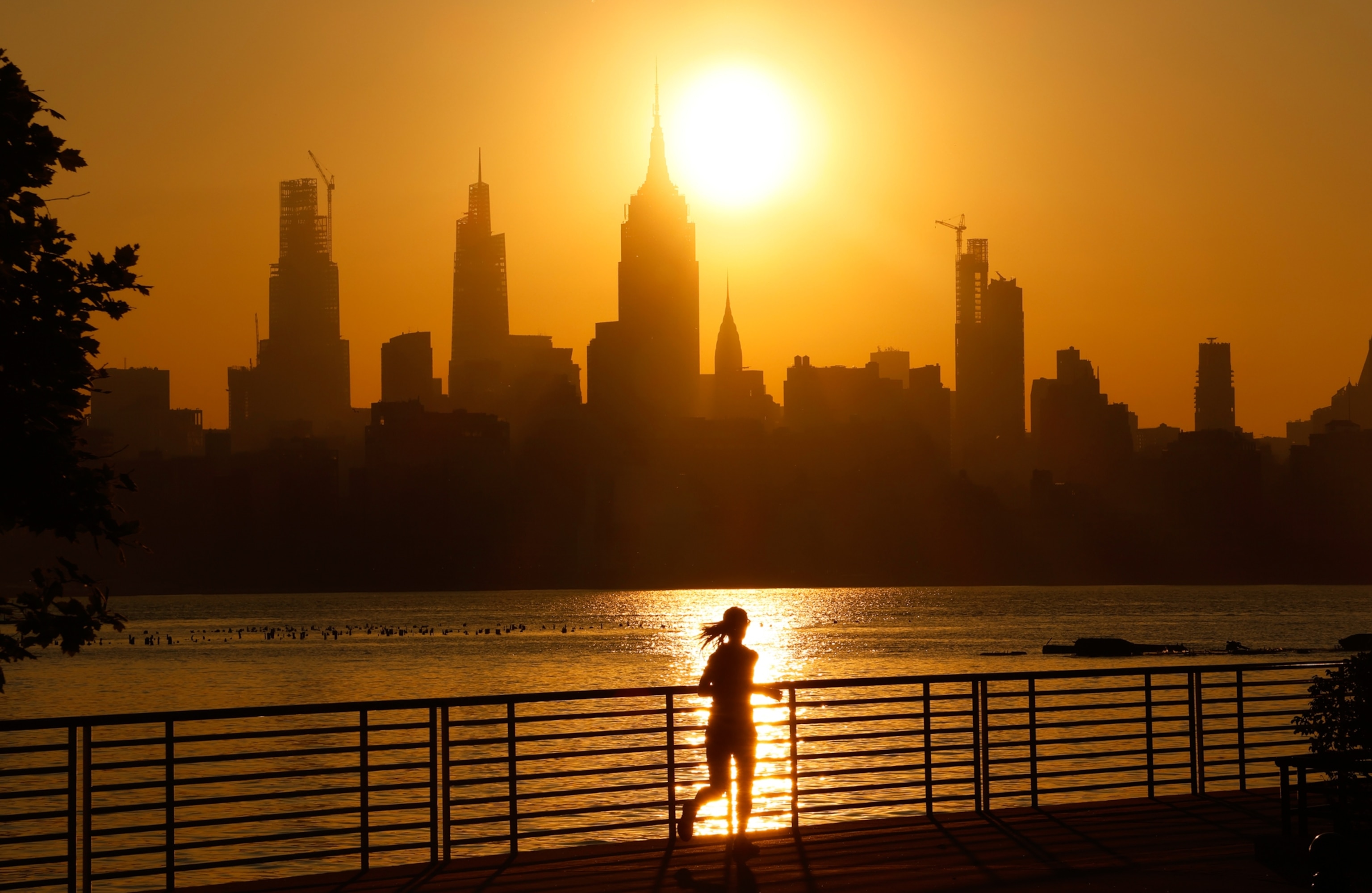 Silhouette of a person jogging along a waterfront at sunrise. The sun casts a warm glow over a city skyline with tall buildings