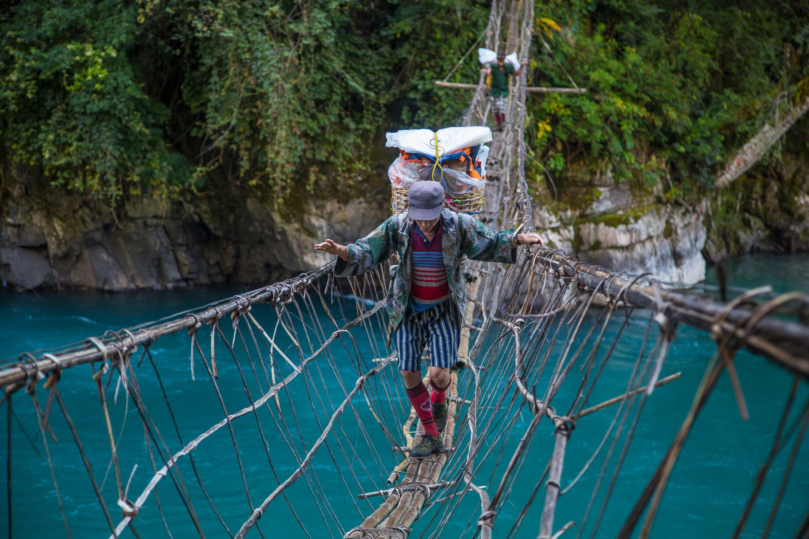 A porter walks on a hanging bridge, which hangs over teal water, as he carries gear for the climbing team that attempted to summit and measure Hkakabo Razi