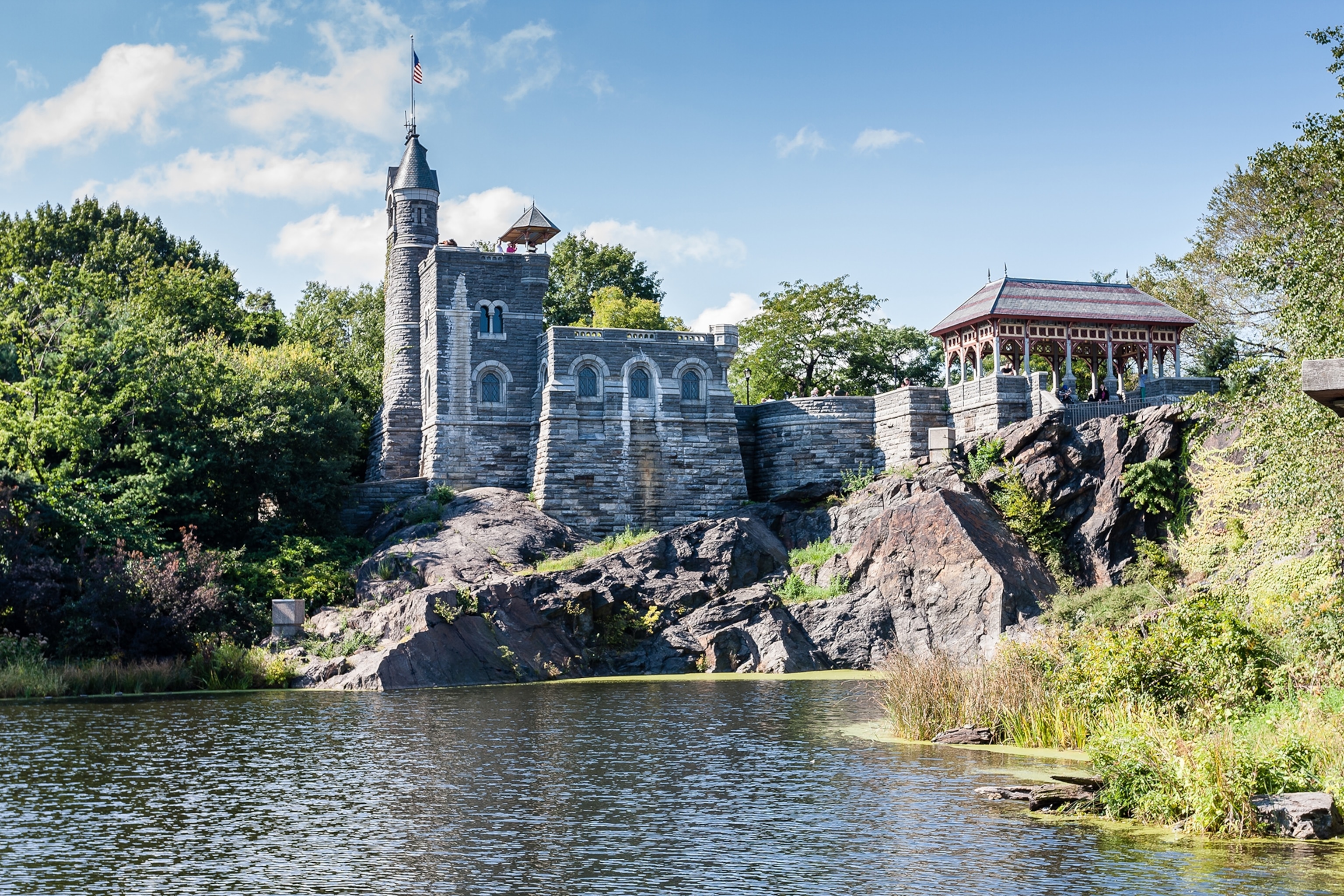 Grey castle with turrets perched on stone surrounded by lush vegetation and a shallow lake in front