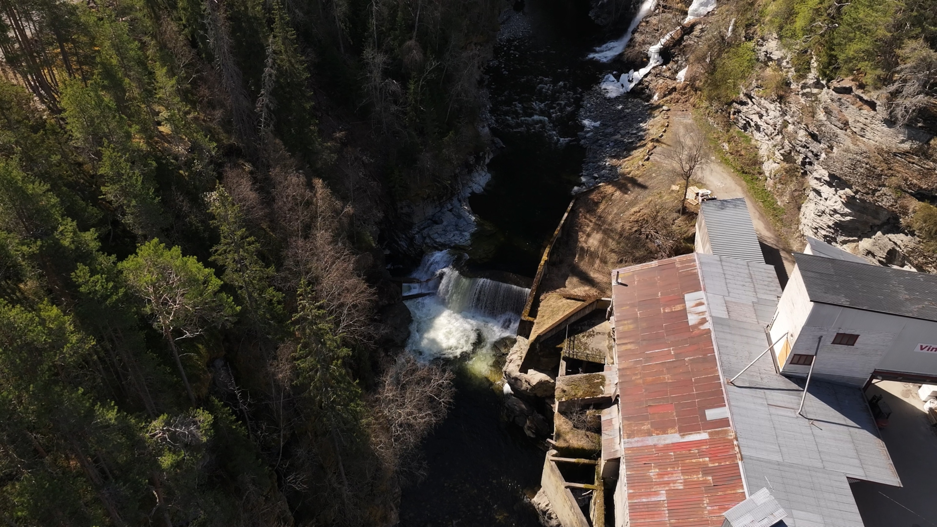 an aerial view of a small waterfall surrounded by dense forest, adjacent to an old industrial building with a rusted roof