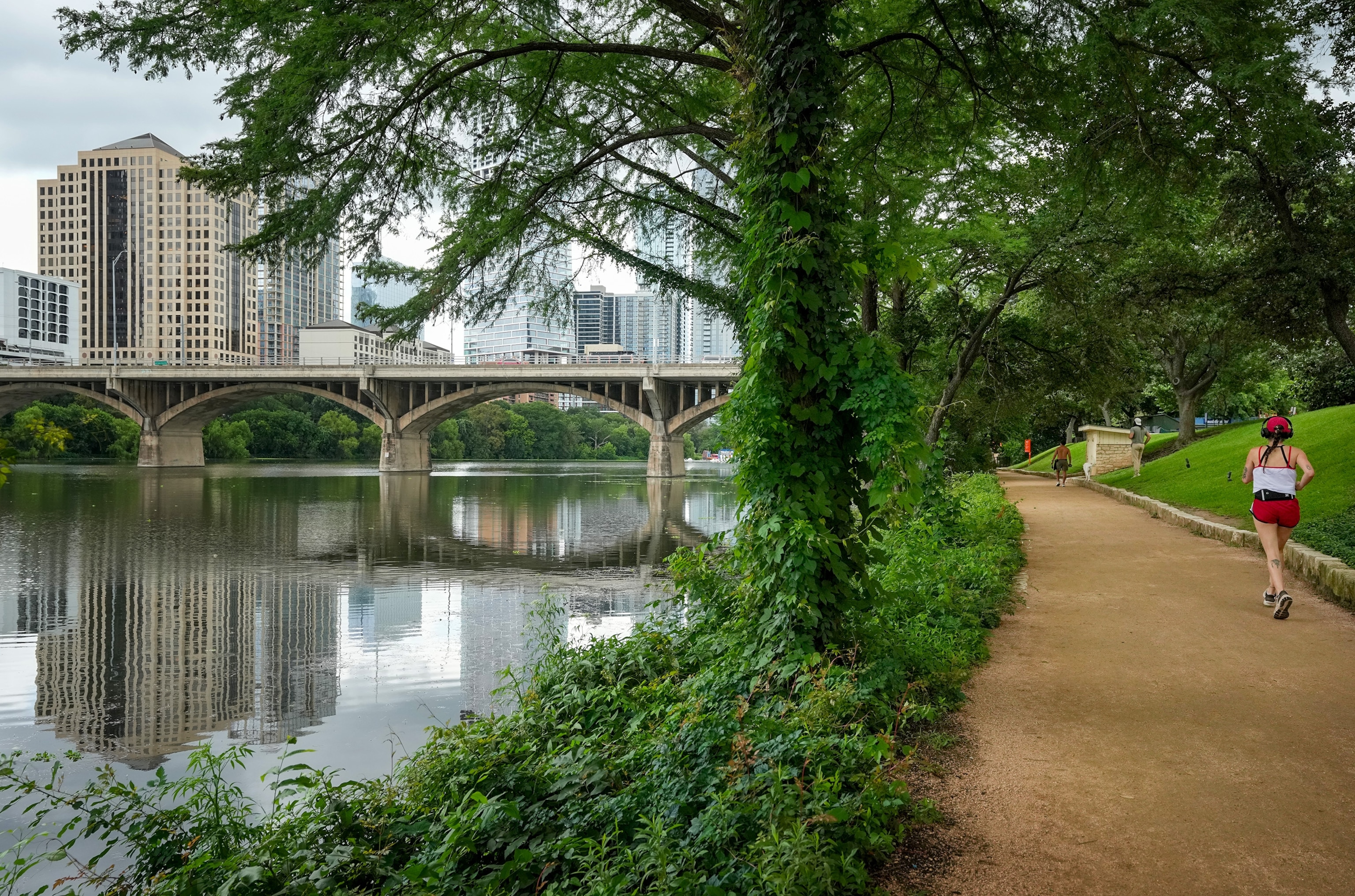 A woman runs down a dirt path alongside a river to the left of her with a bridge and cityscape in the distance.