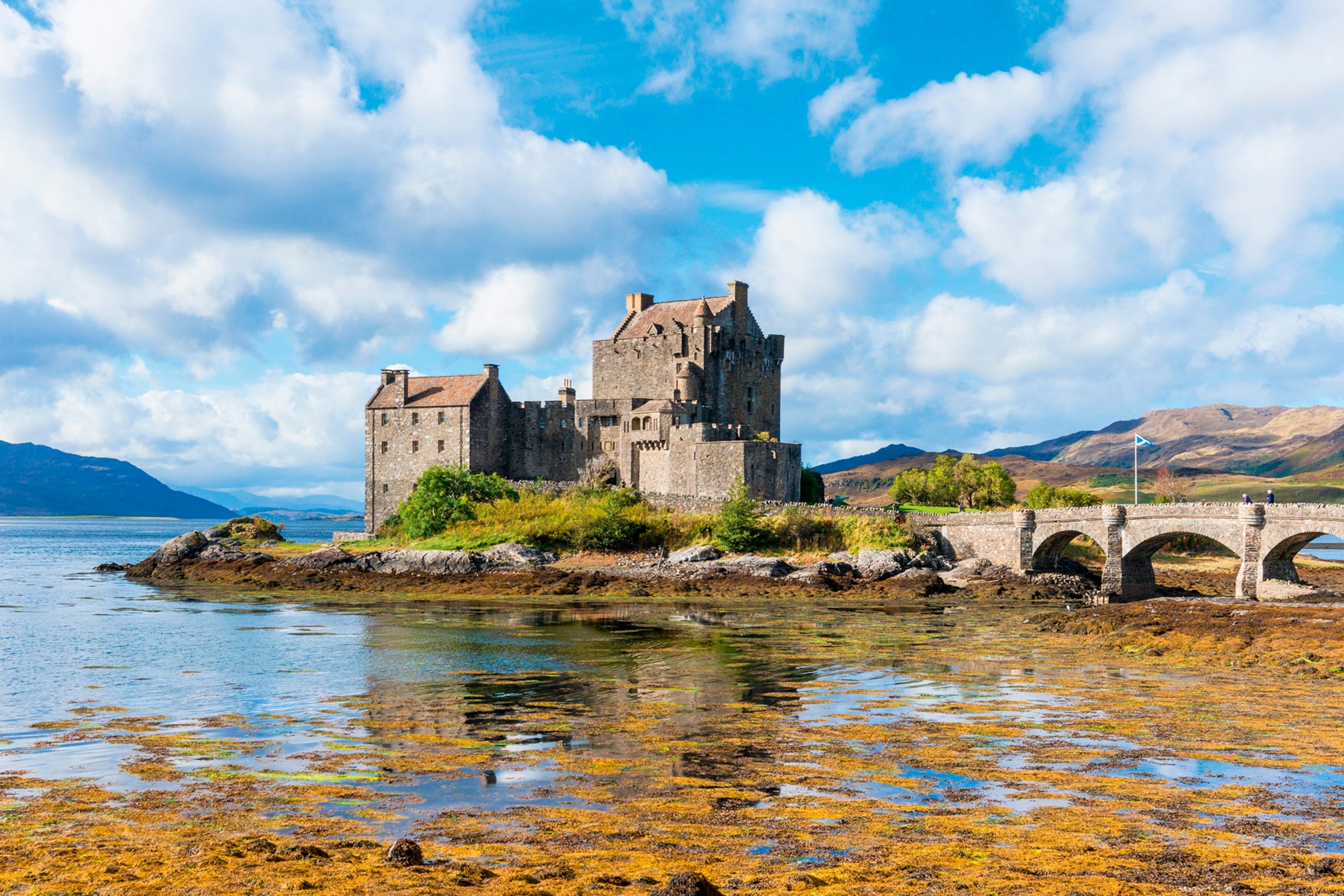 An epic landscape scene of an old, stone castle sitting on the edge of a river with an arched stone bridge leading to its estate.