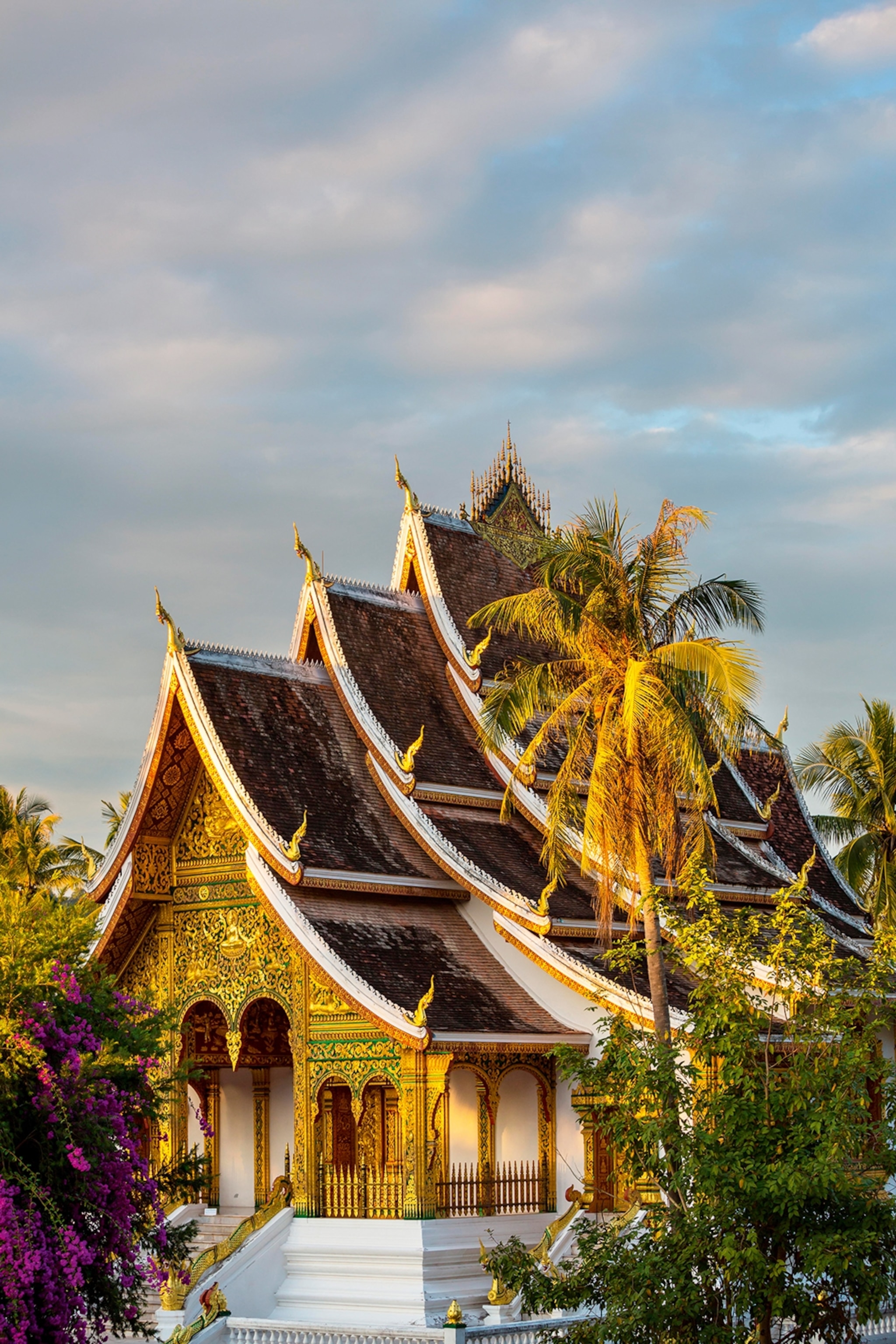 A gold-peaked and multi-tiered temple with an ornate facade in tropical surroundings.
