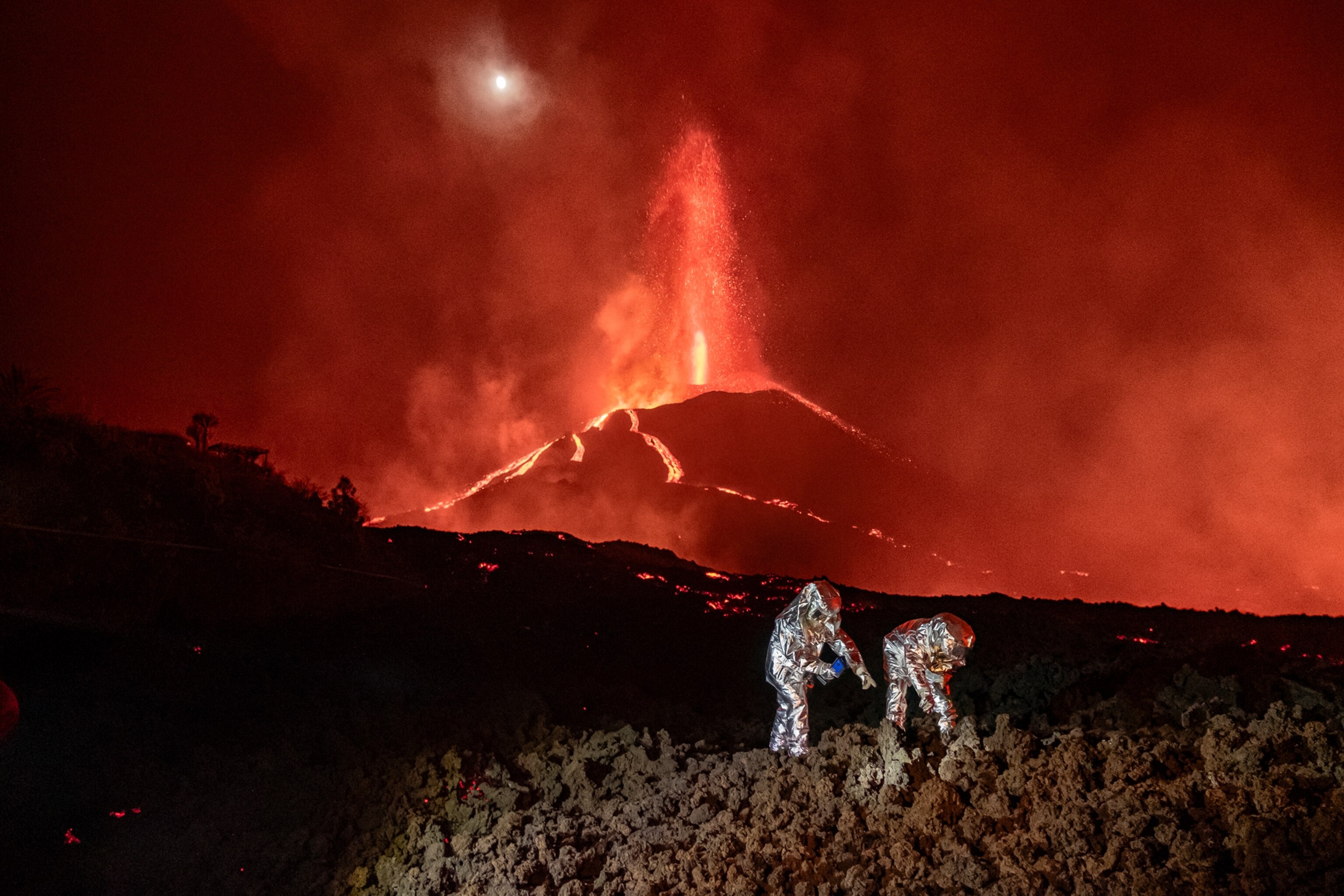 Two scientists wearing silver-colored, full-body protective suits collecting samples from a bed of black volcanic rocks, while a volcano continues to erupt in the background.