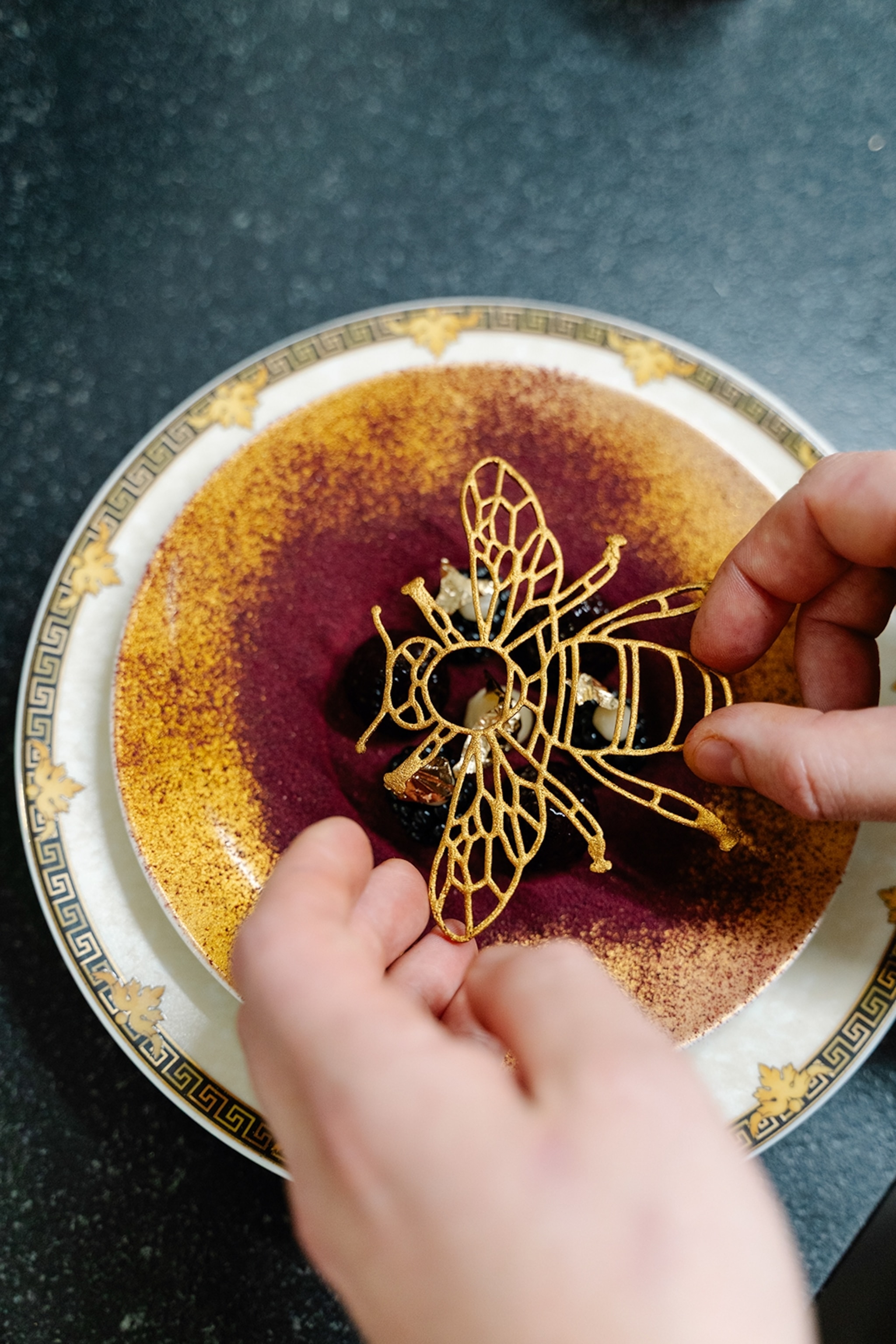 A close-up of two hands placing a delicate chocolate-outlined bee decoration onto the top of a cheesecake.