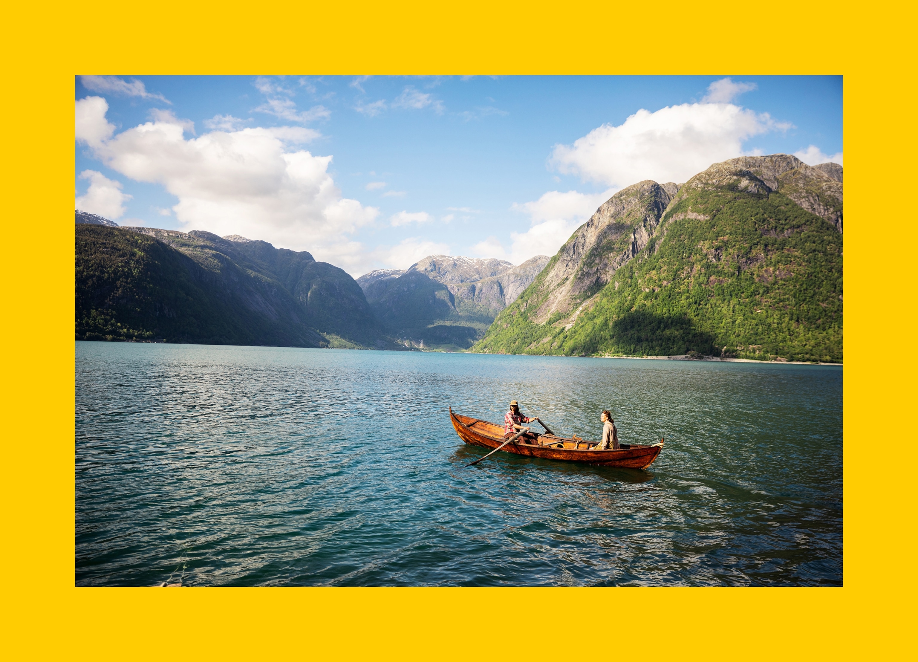 A small wooden rowboat carrying two people floats on a calm fjord, surrounded by steep green mountains and clear blue water under a partly cloudy sky.