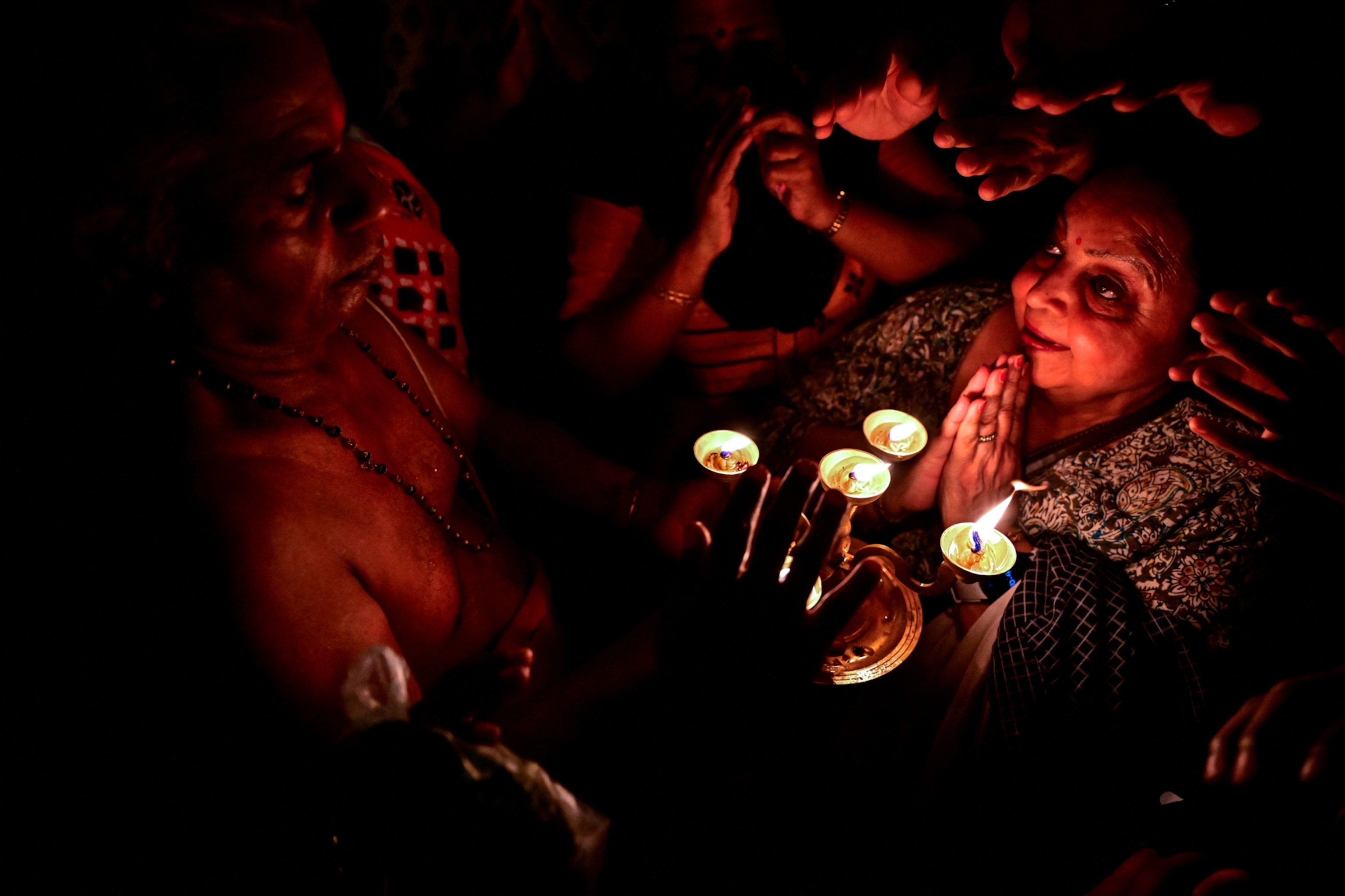 A woman with folded hands looks at a person holding several lit lamp in a dark setting.