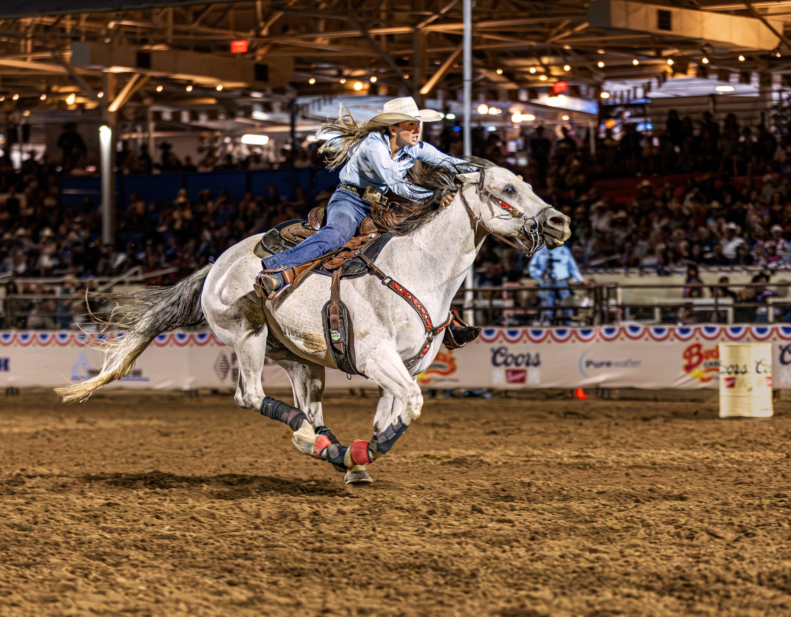 A woman rides a white horse in a dirt arena with a crowd sitting in the stands on the background.