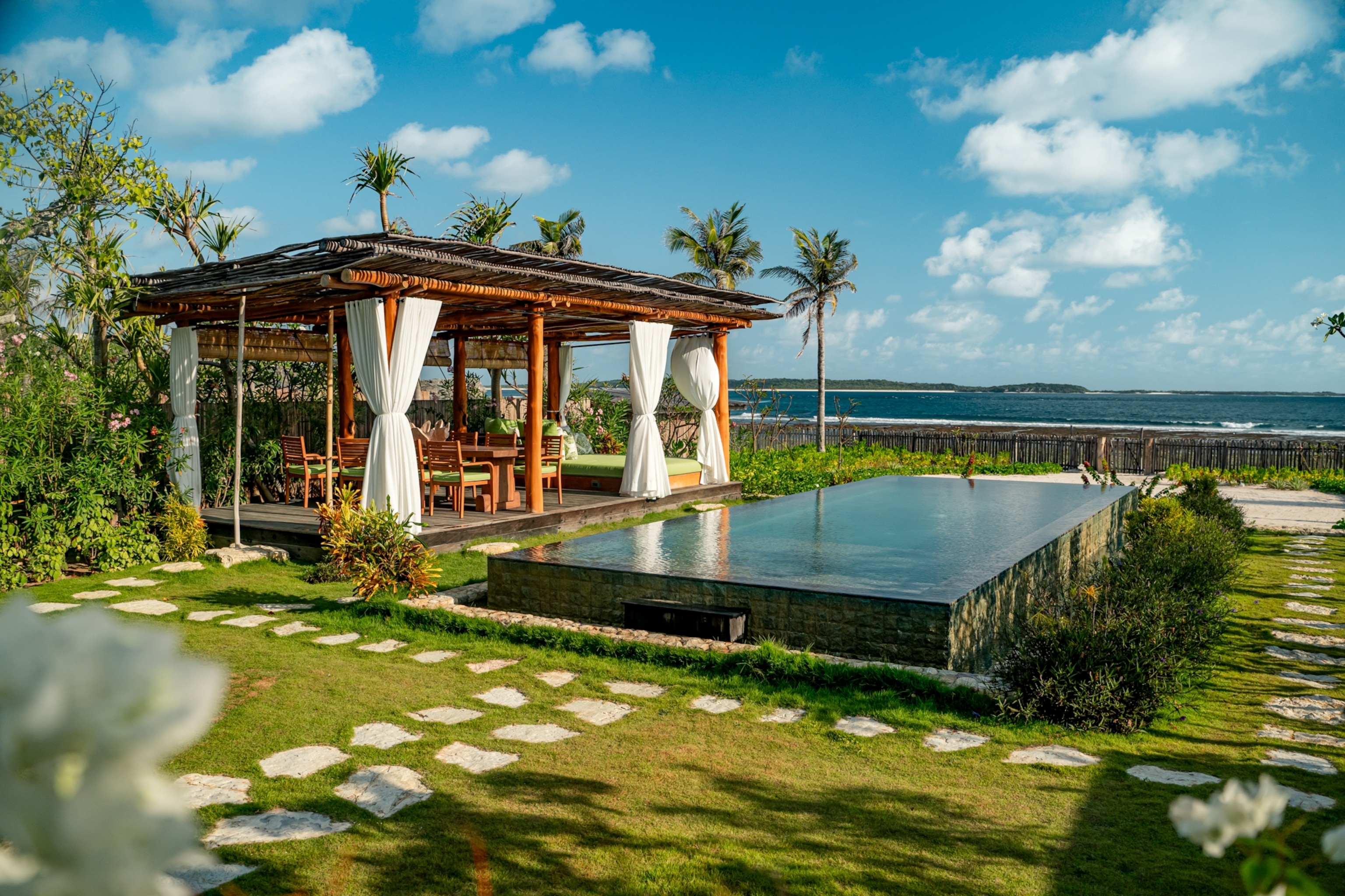 A beachfront villa with an infinity pool, a covered outdoor seating area, palm trees, and views of the ocean under a blue sky.