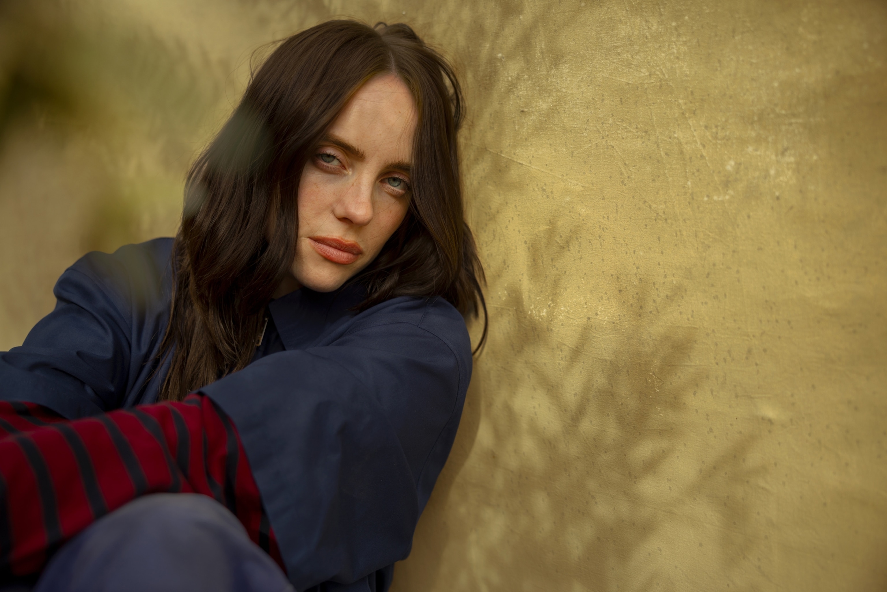 portrait of a woman with dark long hair leaning against a beige wall