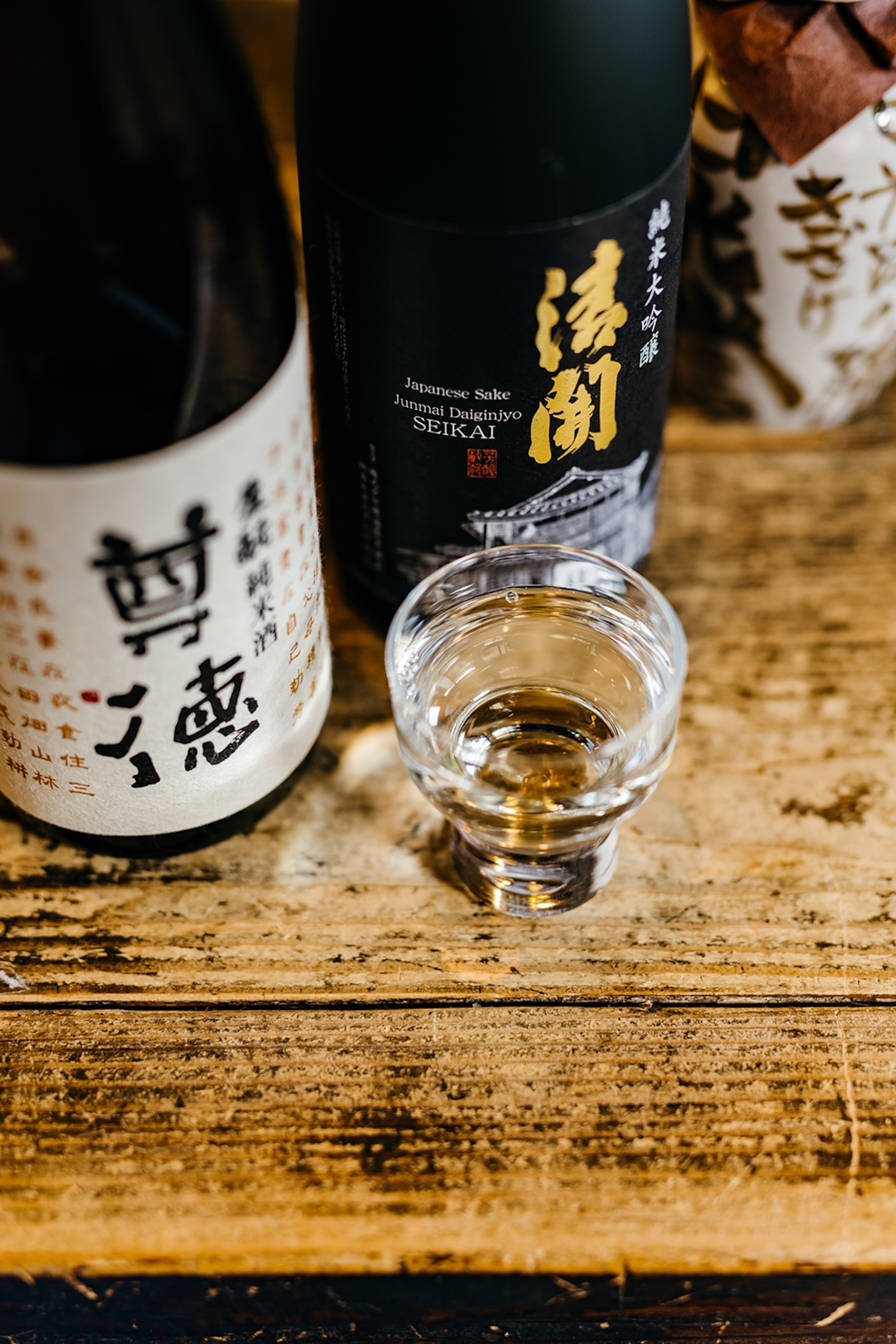 A glass of clear sake next to two bottles on a wooden table.